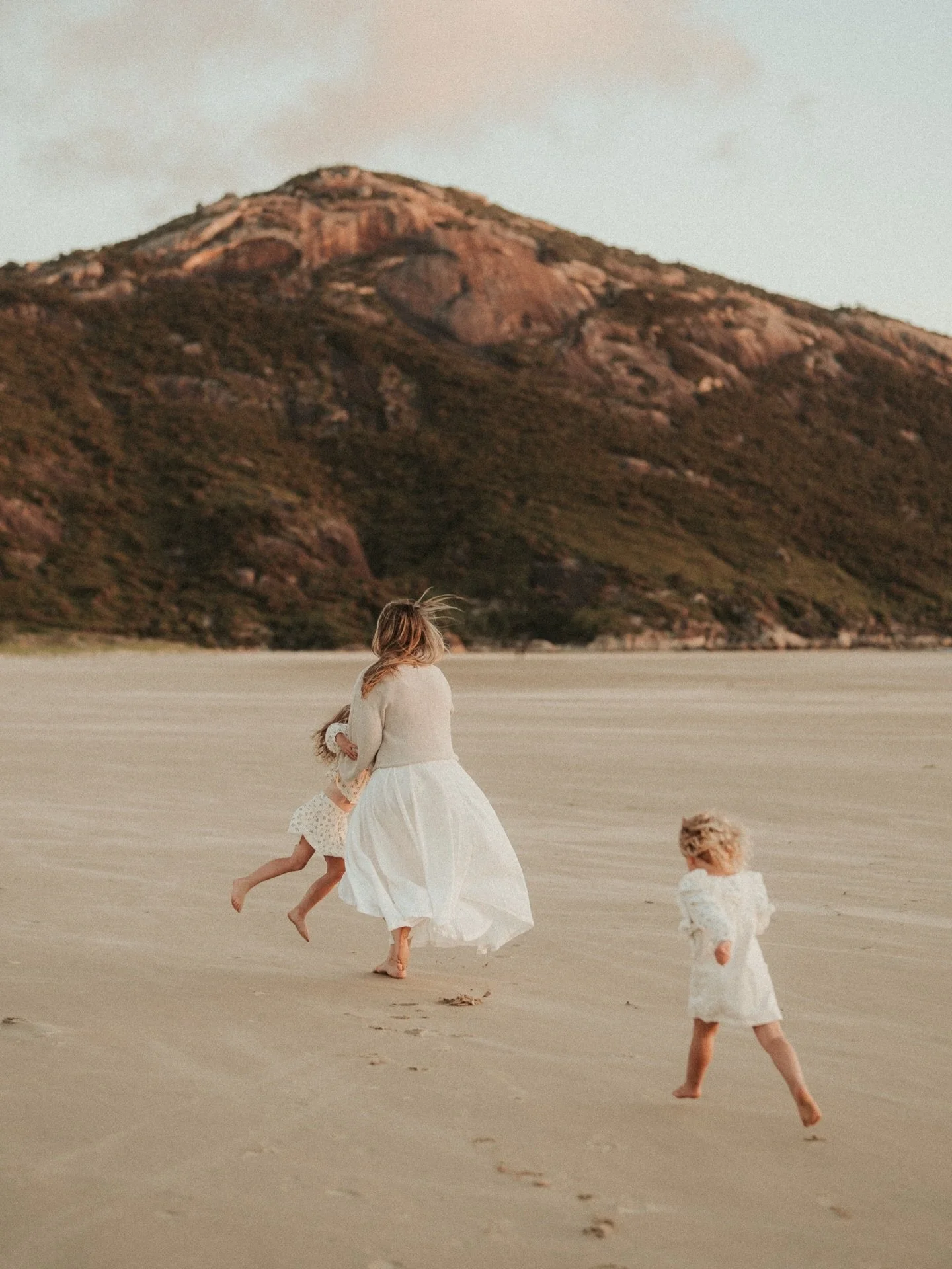 Three children and a woman playing and running on a sandy beach with mountains in the background during sunset.