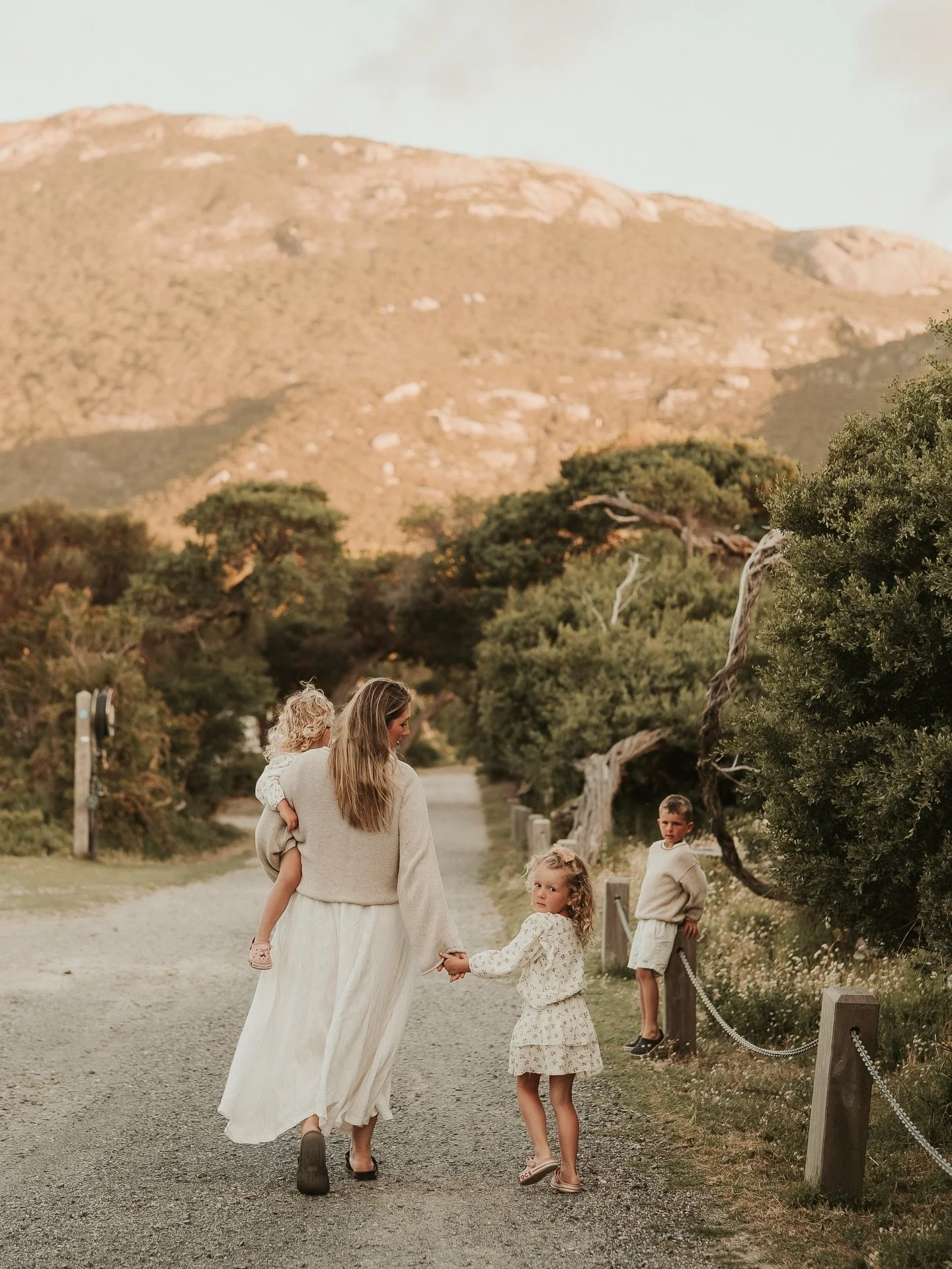 A woman holding a young girl by the hand walking along a gravel path with three other children nearby, surrounded by trees and a mountain in the background during sunset.