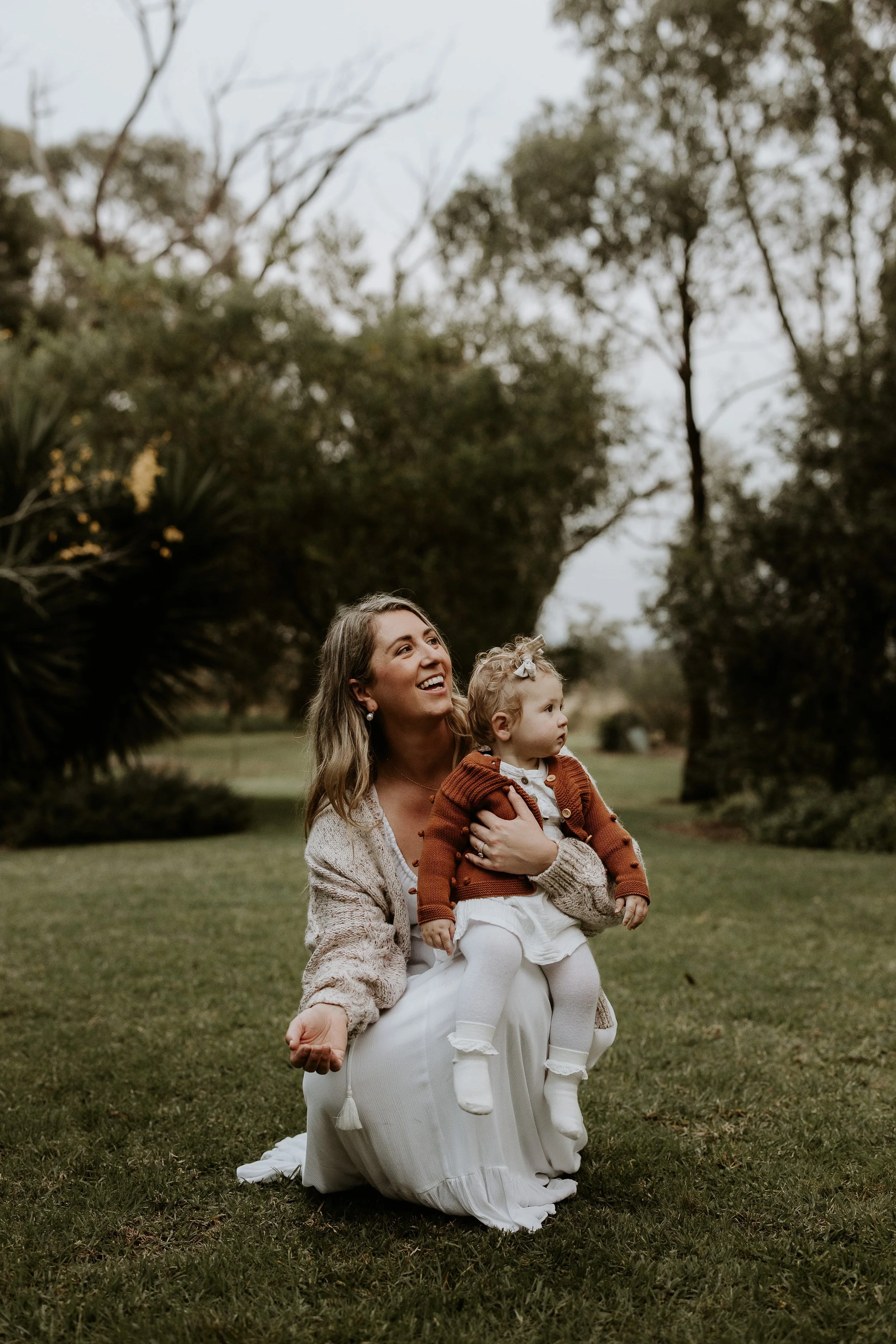 A woman kneeling on the grass outside, holding a young girl dressed in white, with trees and cloudy sky in the background.