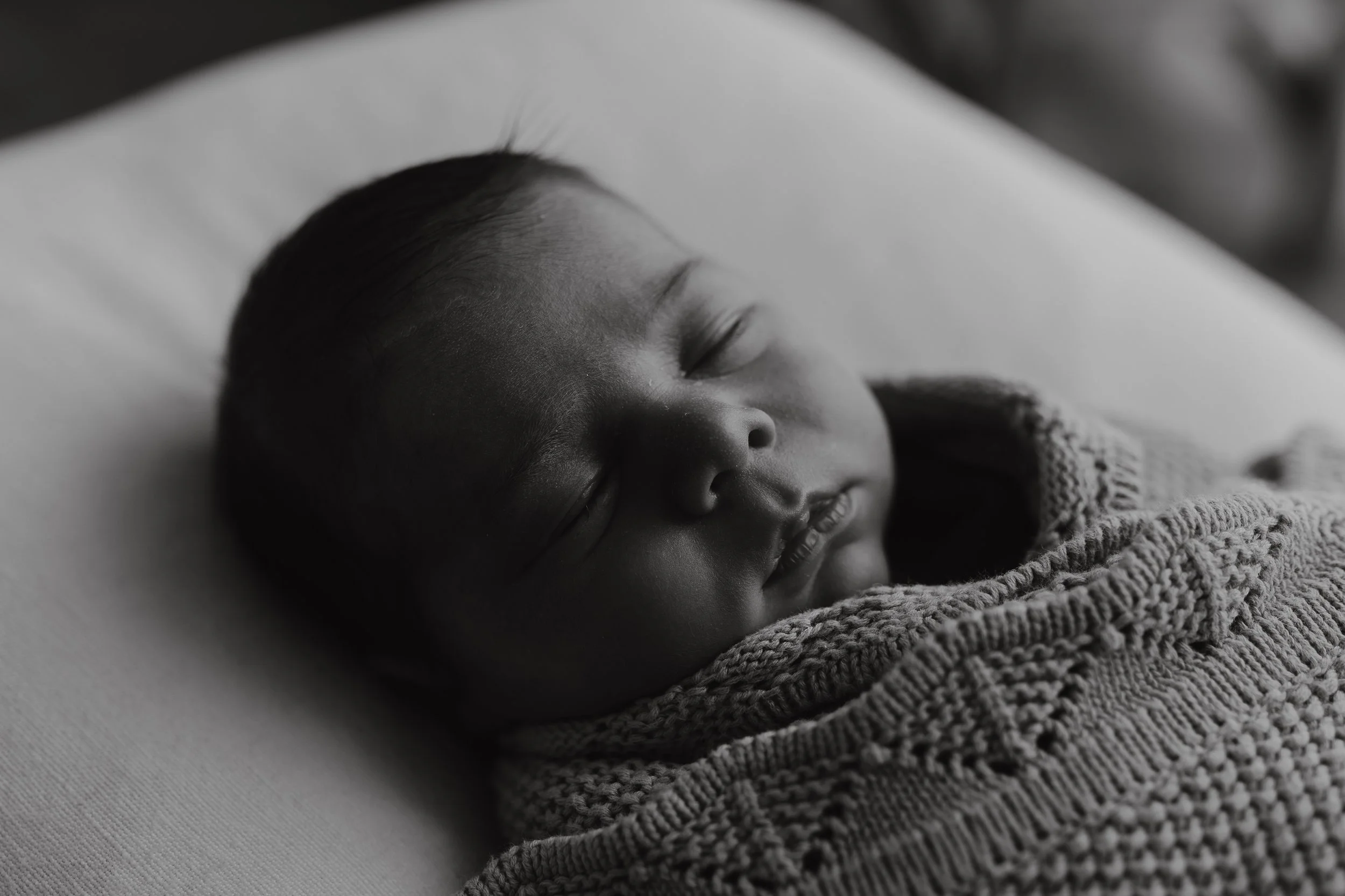 Close-up of a baby wearing a beige knitted sweater, lying on a dark surface, with eyes partially open and hands near the face.