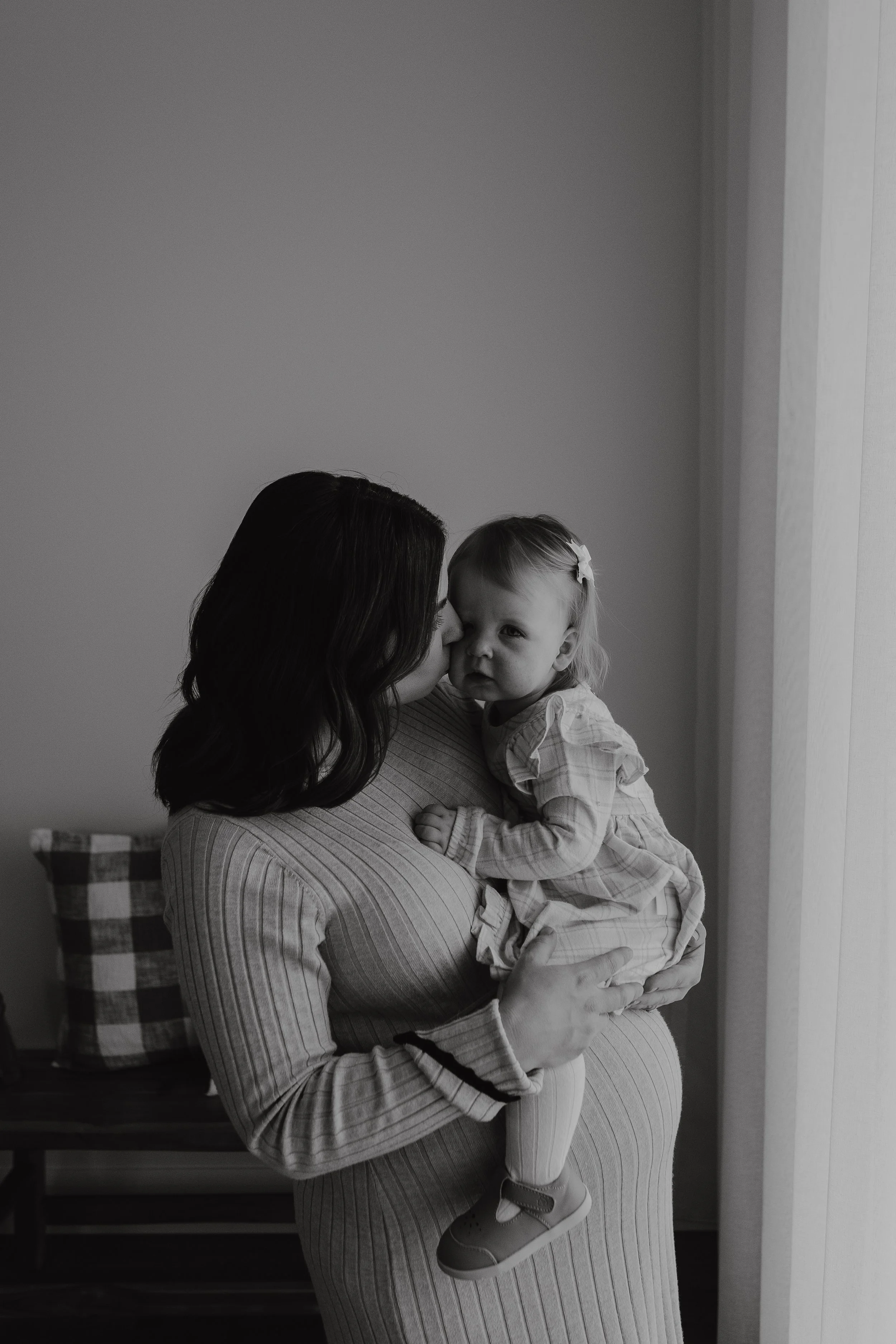  A black and white photo of a woman with medium length, wavy hair is standing holding and looking at  a young child who is wearing a dress, in a cozy bedroom style studio with neutral-toned colours and pillows.
