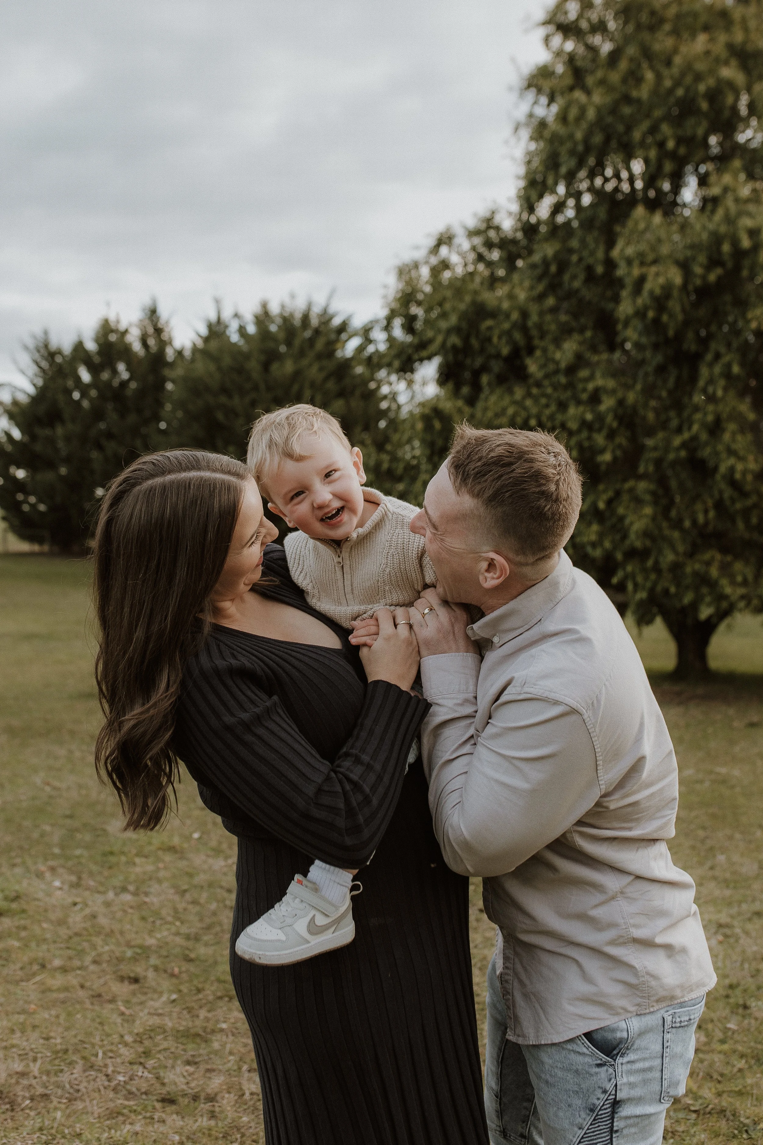 A pregnant woman and a man both smiling at and holding a small boy in-between them, as he laughs in a grassed outdoor field with trees in the background. Woman is wearing a dark knit dress, the man is wearing a beige shirt and jeans, and the small bo