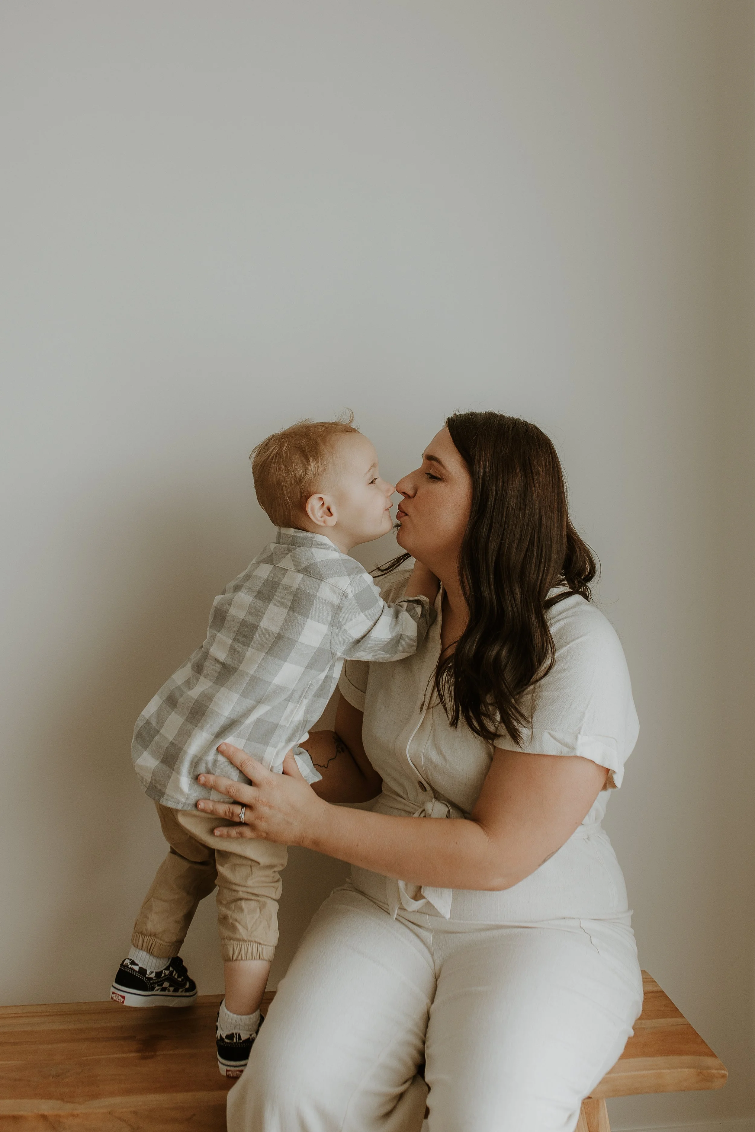 A woman with long, brown wavy hair is sitting on a bench, holding and kissing a young child who is wearing a checked shirt and beige pants, in a cozy bedroom styled studio with neutral-tones