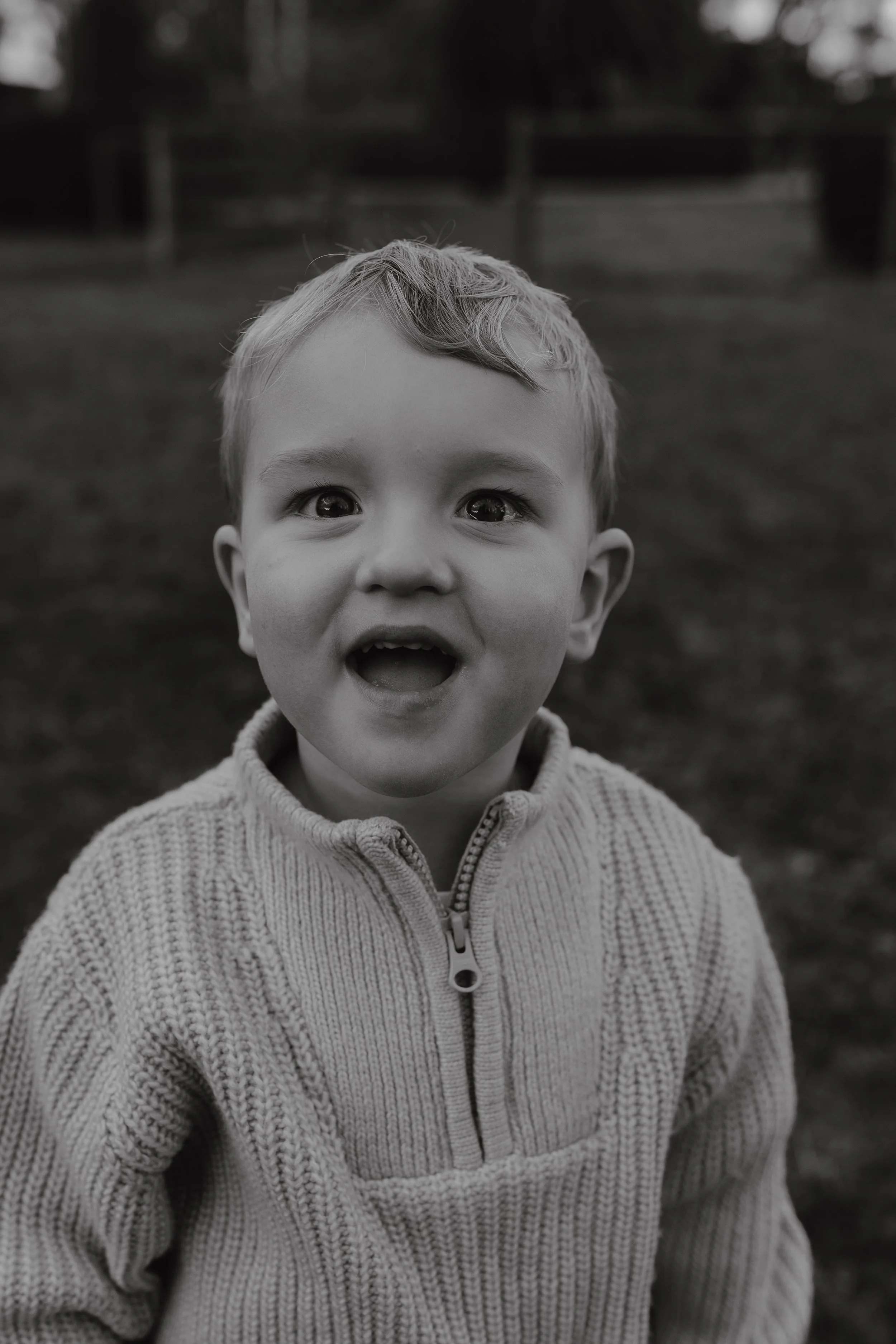 A black and white image of a young boy looking happy and smiling at the camera, wearing a light coloured zip up knitted jumper