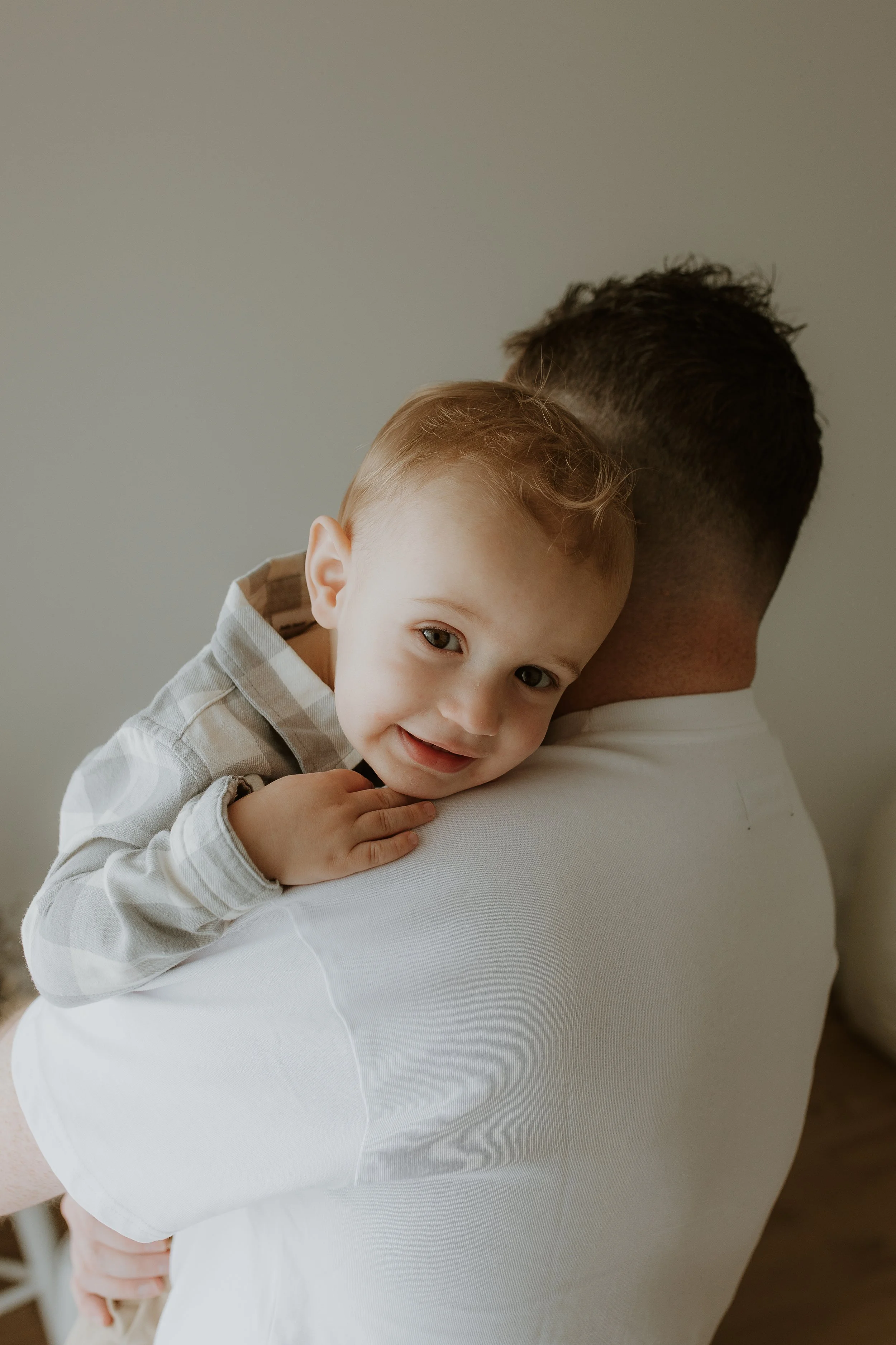 A man with brown hair holding a smiling young boy indoors in a studio, with the child hugging over his shoulder.