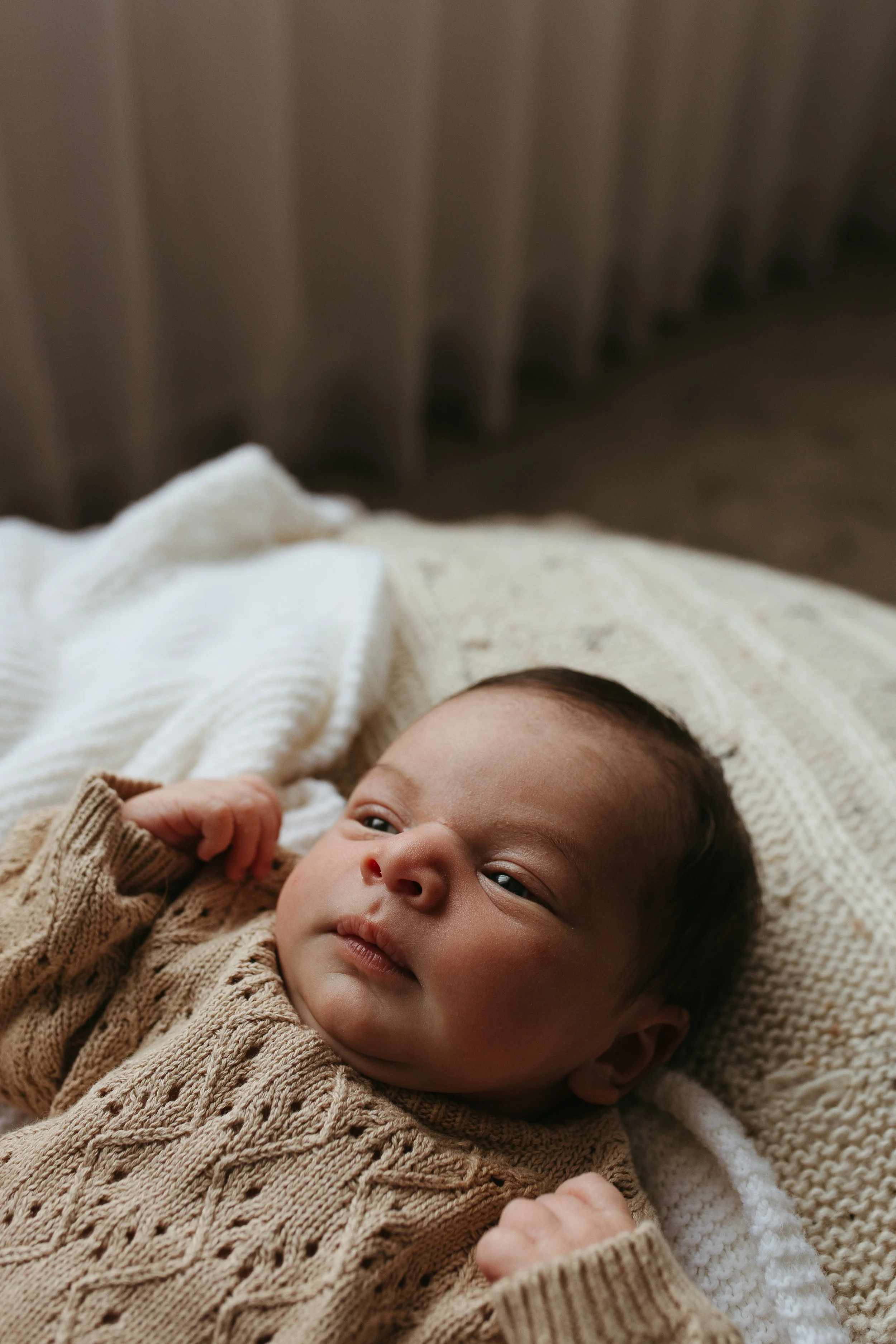 A young baby lying down looking up, with a neutral knitted jumper on, in a room with soft neutral tones