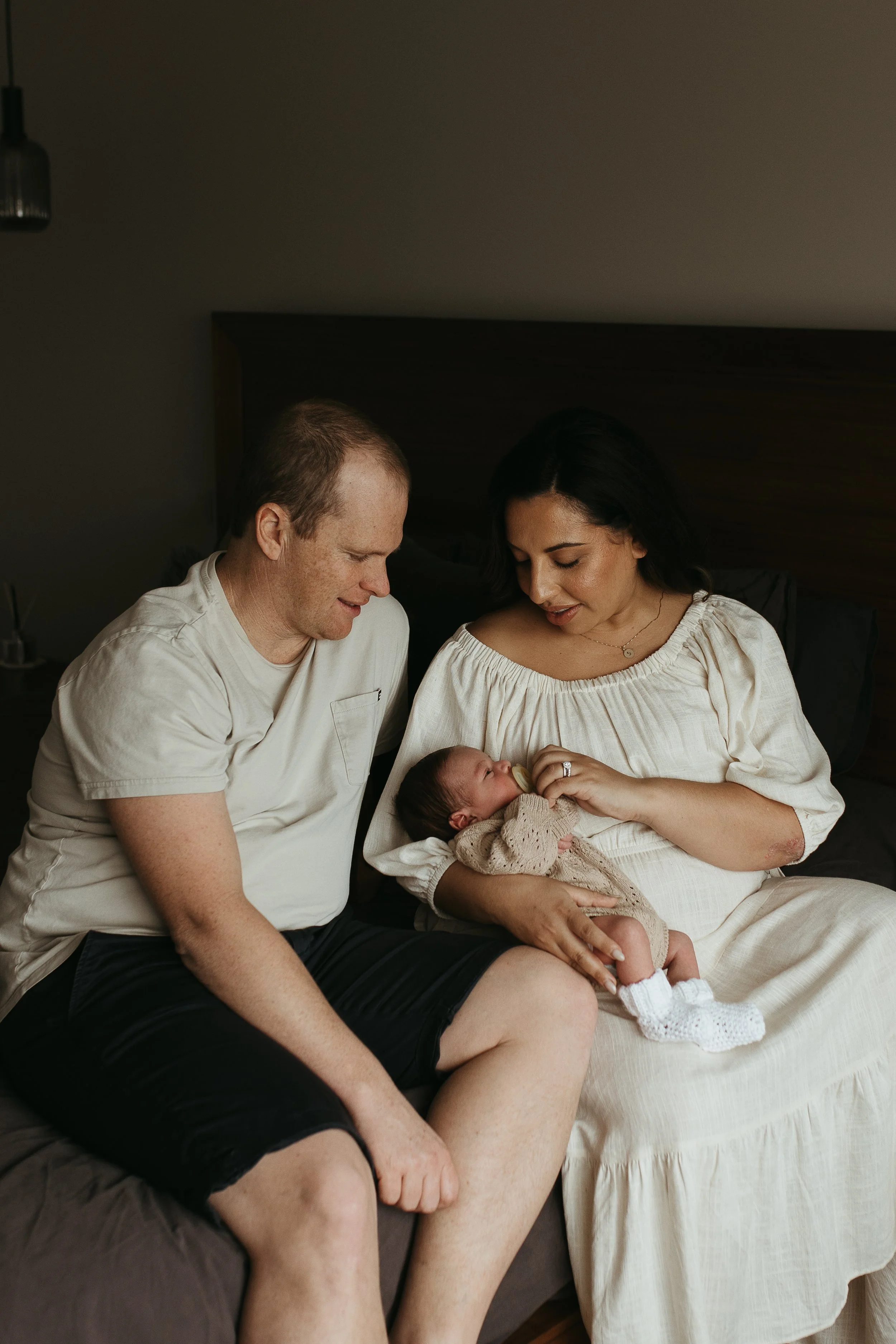 A man wearing dark shorts and a beige top is sitting on a bed with a woman in a beige dress, both looking down and smiling at a small baby in her arms.