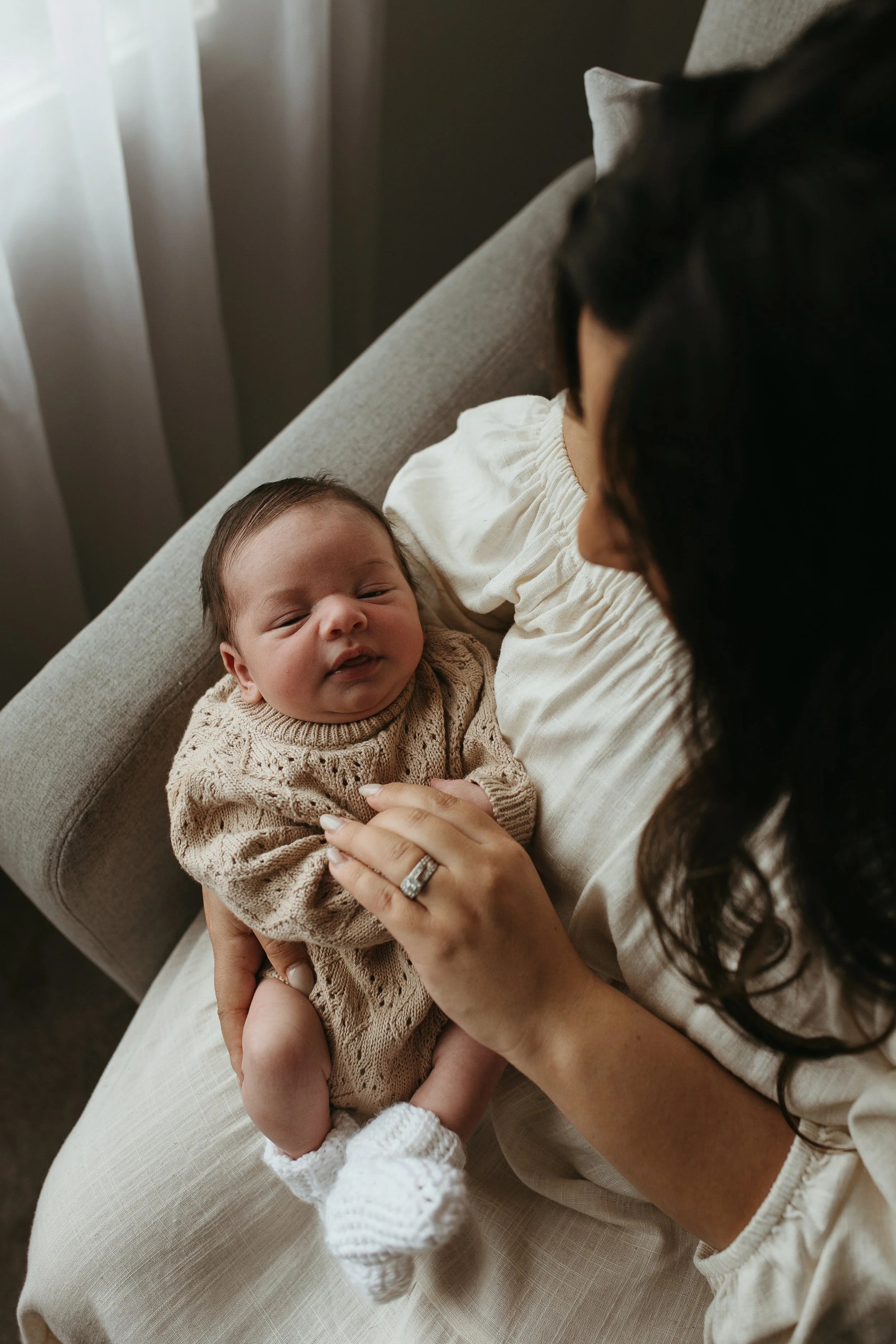 A woman with long, dark hair is sitting on a chair, holding and looking at a young baby who is wearing a beige sweater, in a cozy bedroom with neutral-toned bedding and pillows.