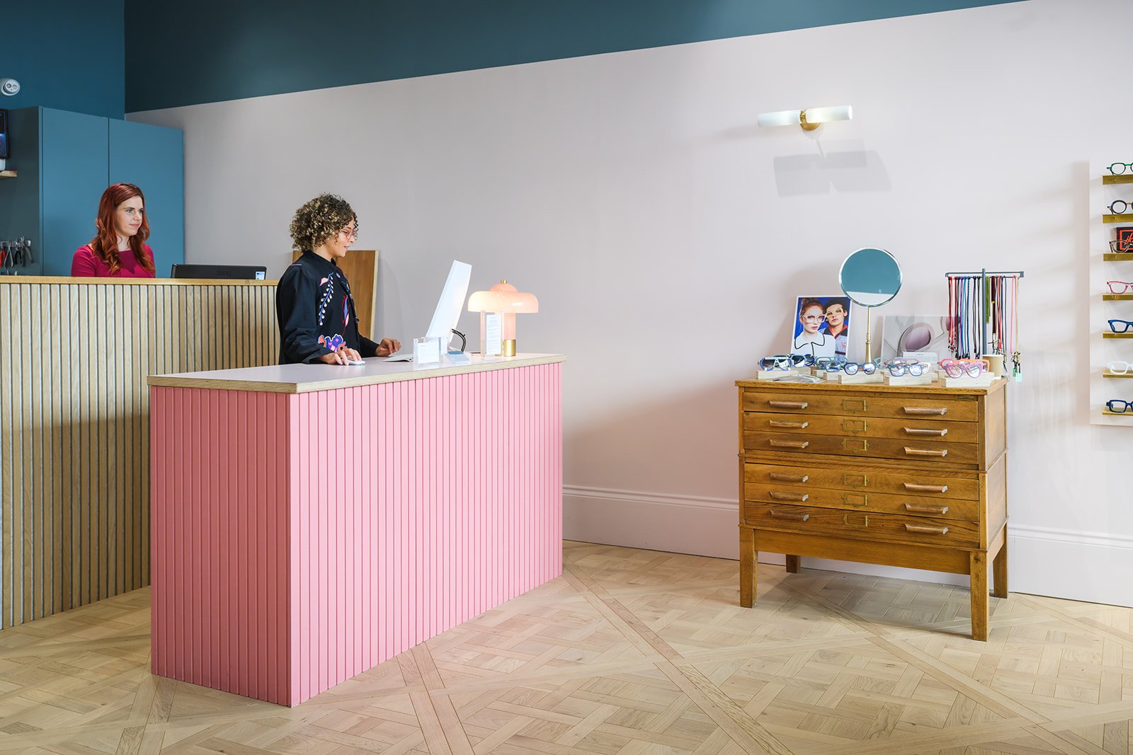 A boutique optometry practice interior with a counter on the left, two women behind each counter, with pink and blue fixtures, and a wooden display table on the right showcasing glasses and accessories.