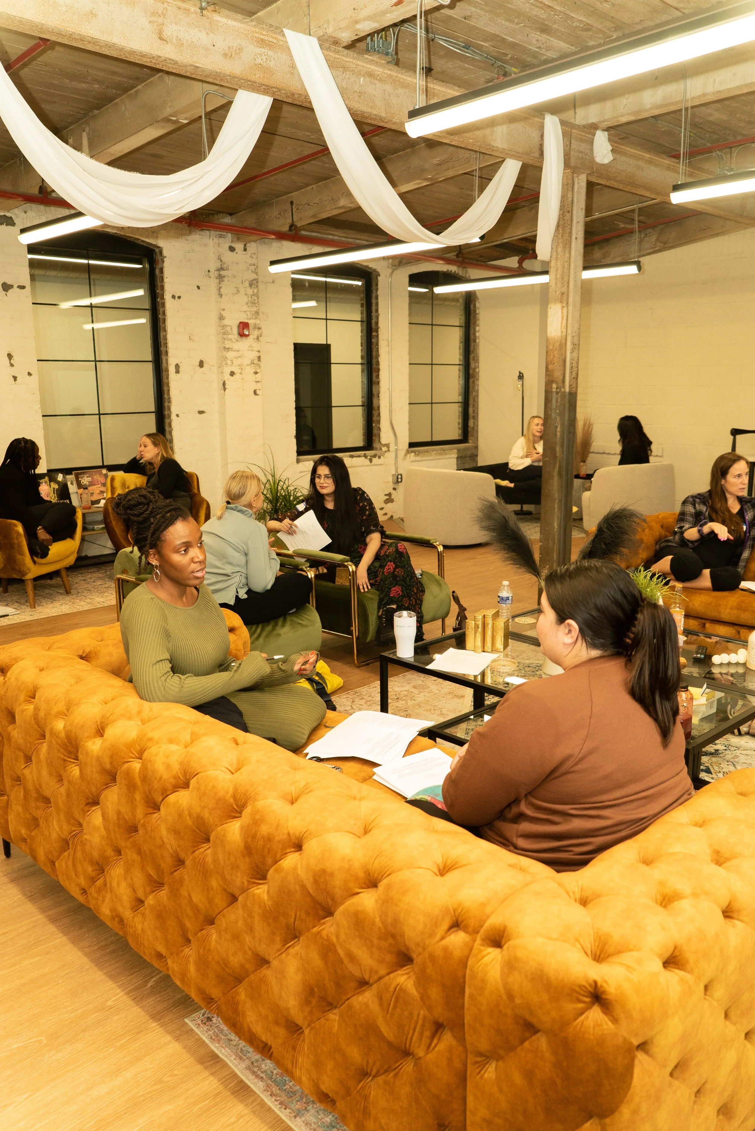 People sitting and talking in a cozy, modern lounge with tufted orange sofas, green chairs, and large windows in an industrial-style space.