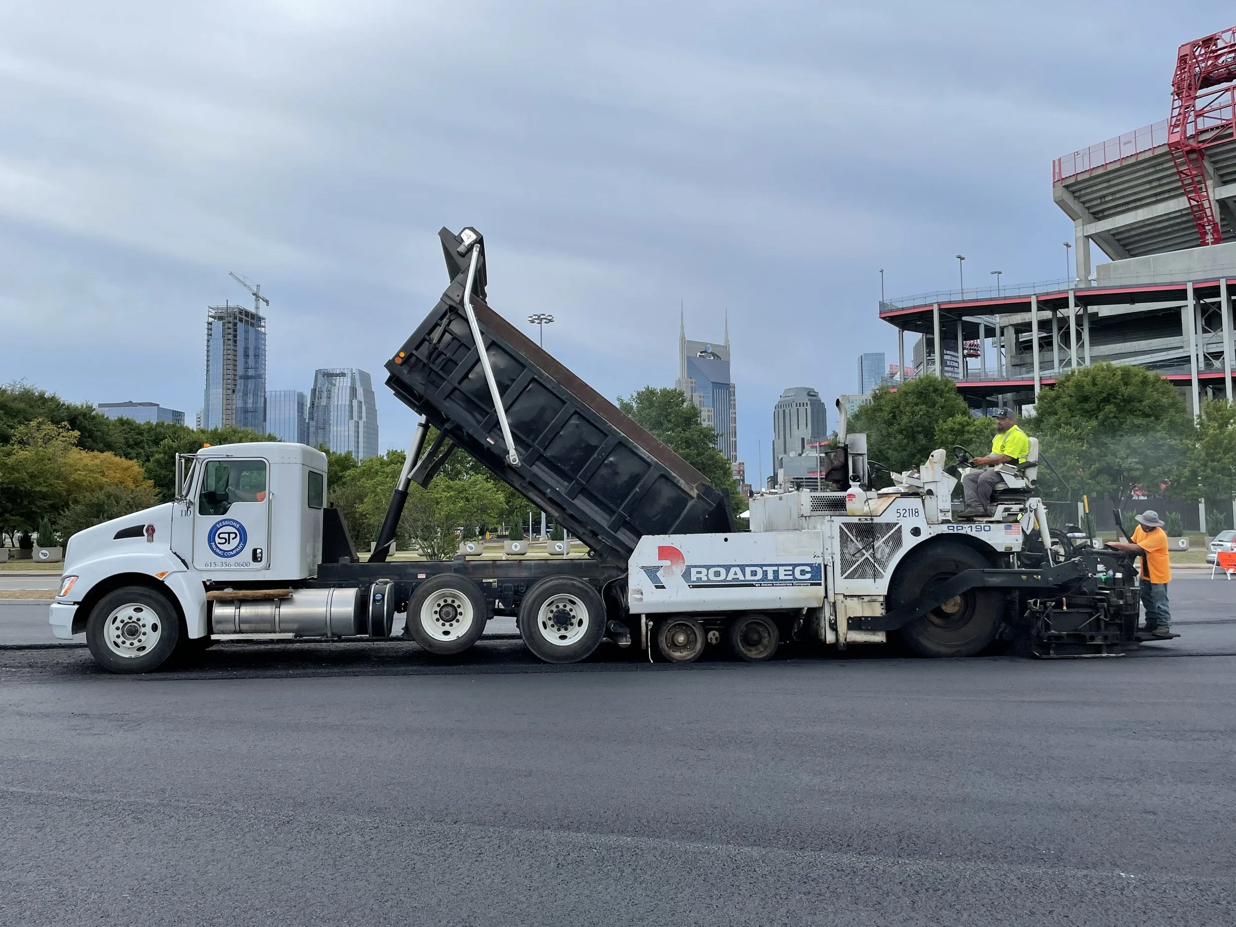 A dump truck, paver, and crew paving asphalt road (pit lane) in parking lot of Nissan Stadium in Nashville, Davidson County, TN for the Music City Grand Prix project using asphalt surface