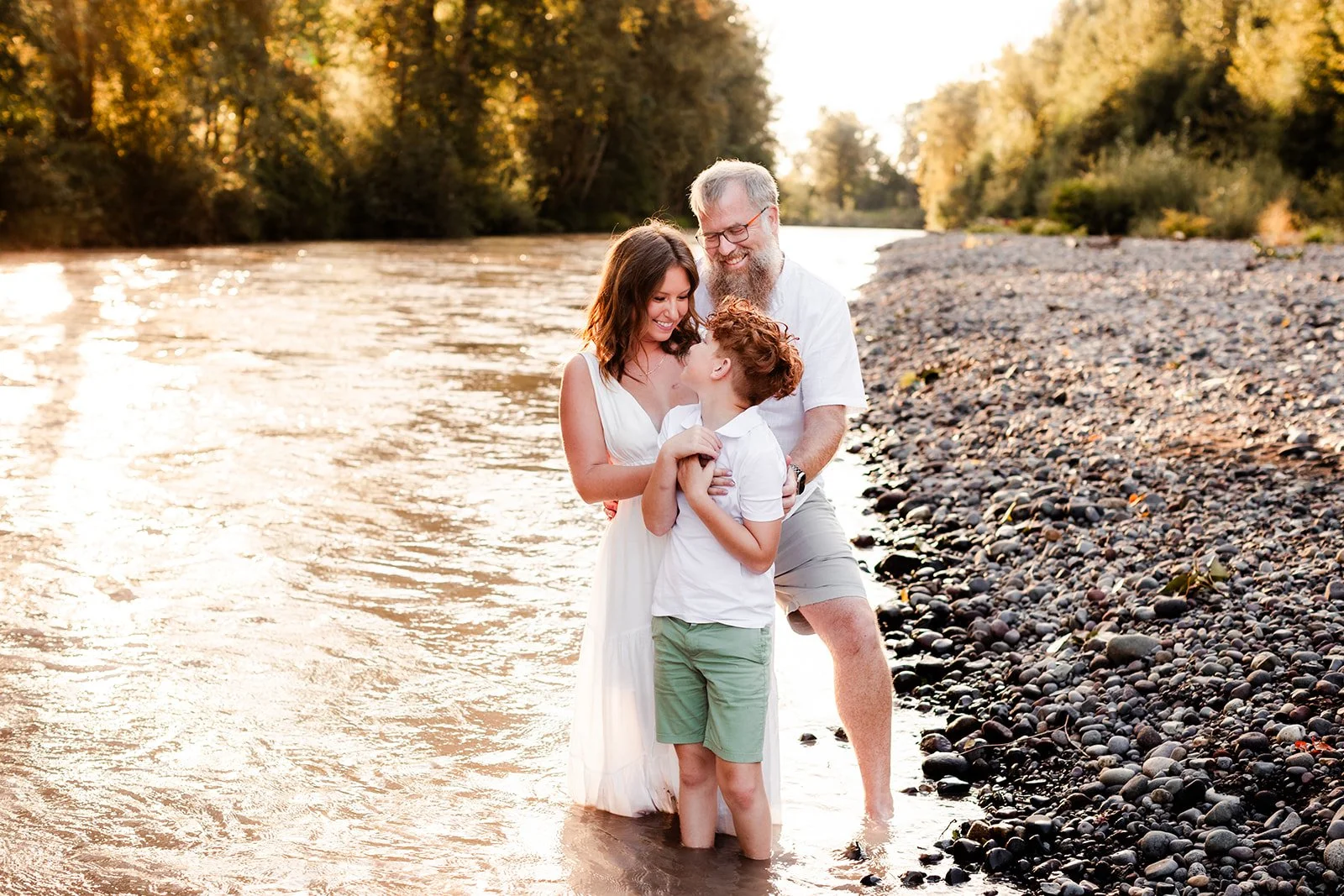 Family of 3 are by a river smiling at each other