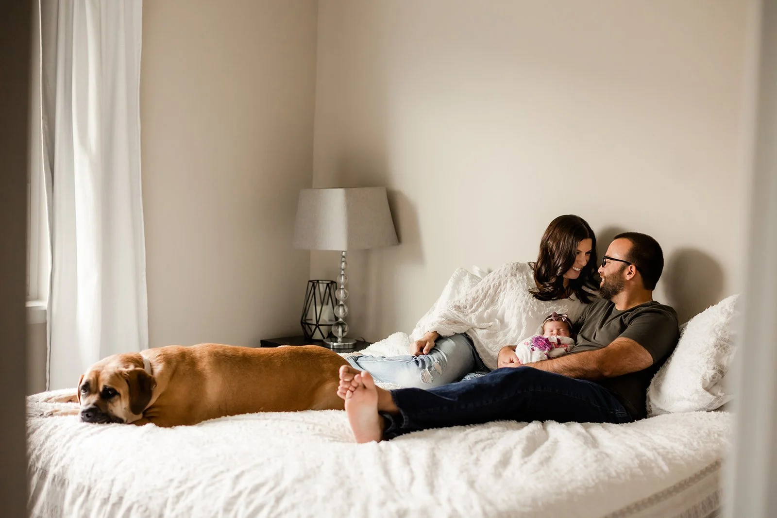 mom and dad sit on bed with newborn baby in arms and dog at their feet