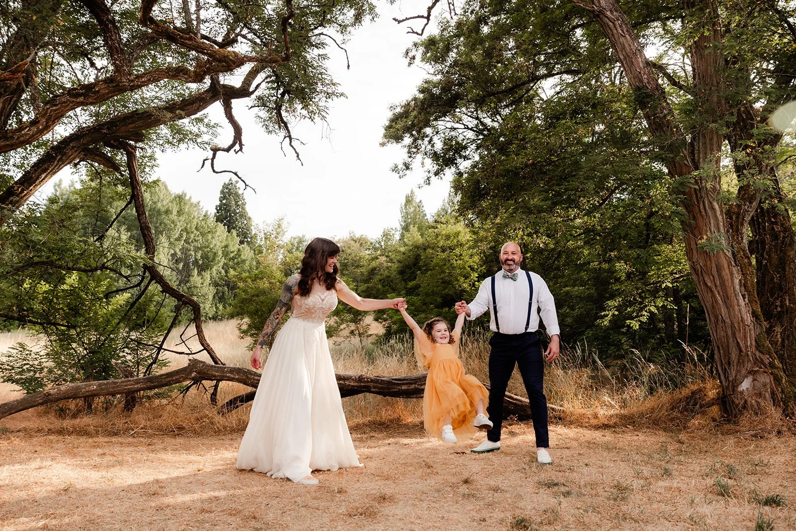 Mom and dad happily swing little girl with oak trees in background