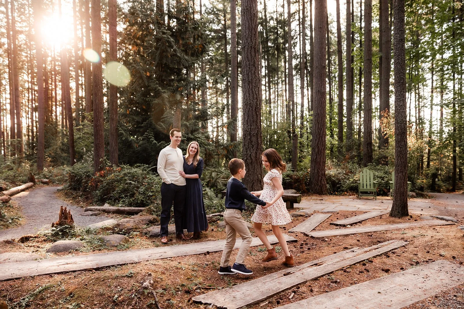 Brother and sister play in the forest while parents smile on at them