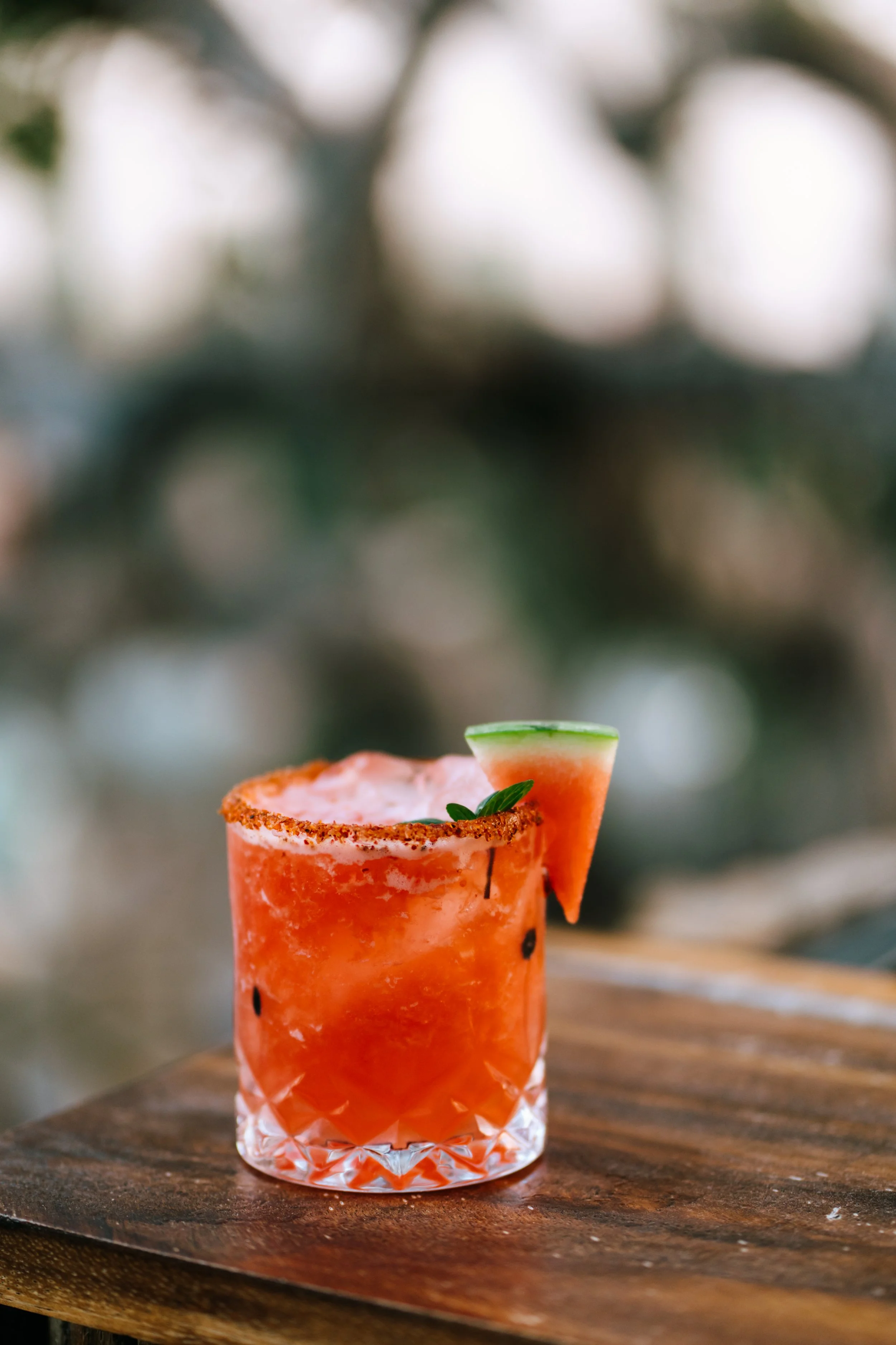 A pink cocktail with a salted rim, garnished with a small watermelon wedge and a mint leaf, served in a textured glass on a wooden surface.