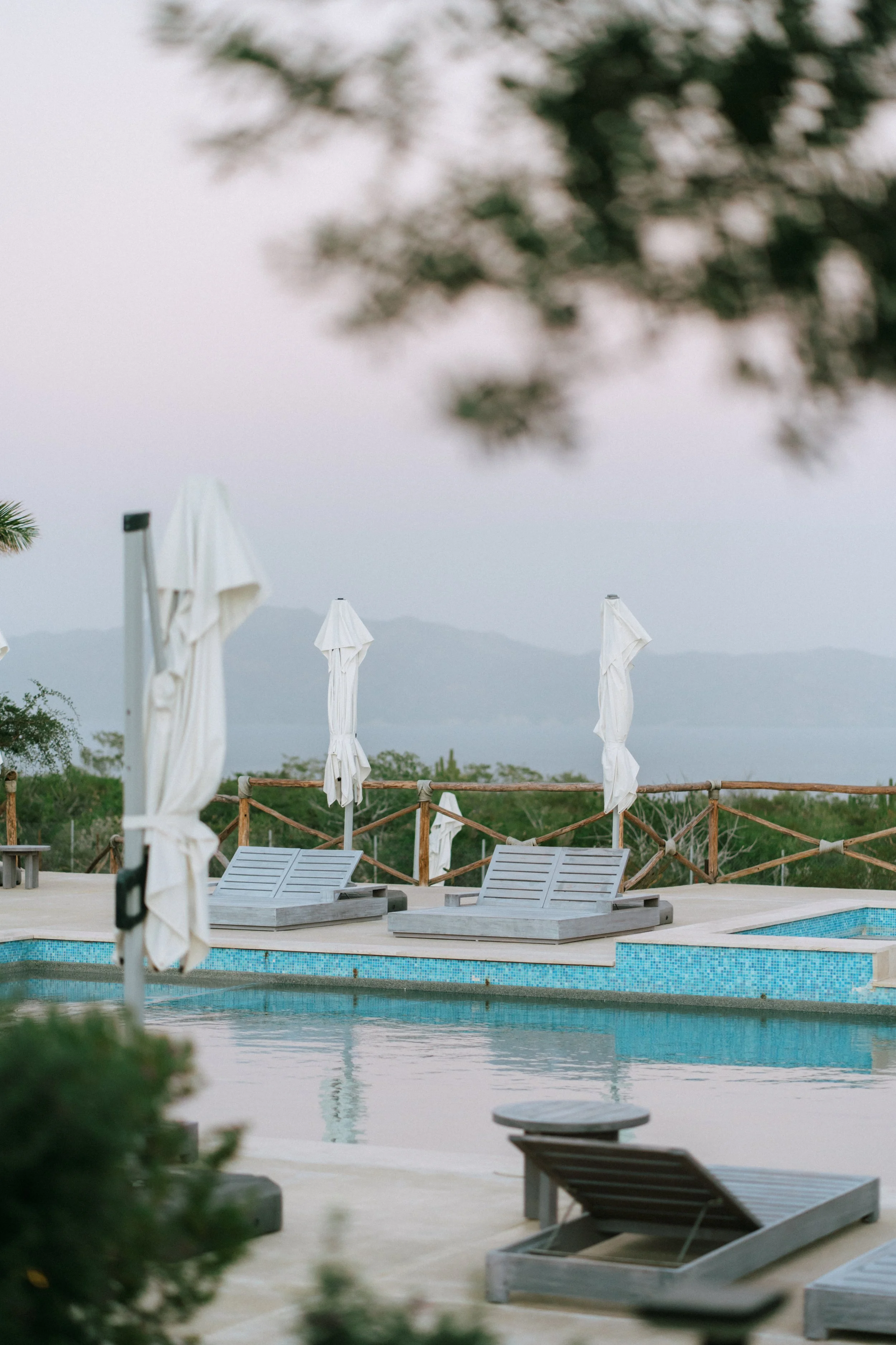 Empty poolside area with lounge chairs and umbrellas, overlooking a distant body of water and mountains under a cloudy sky.