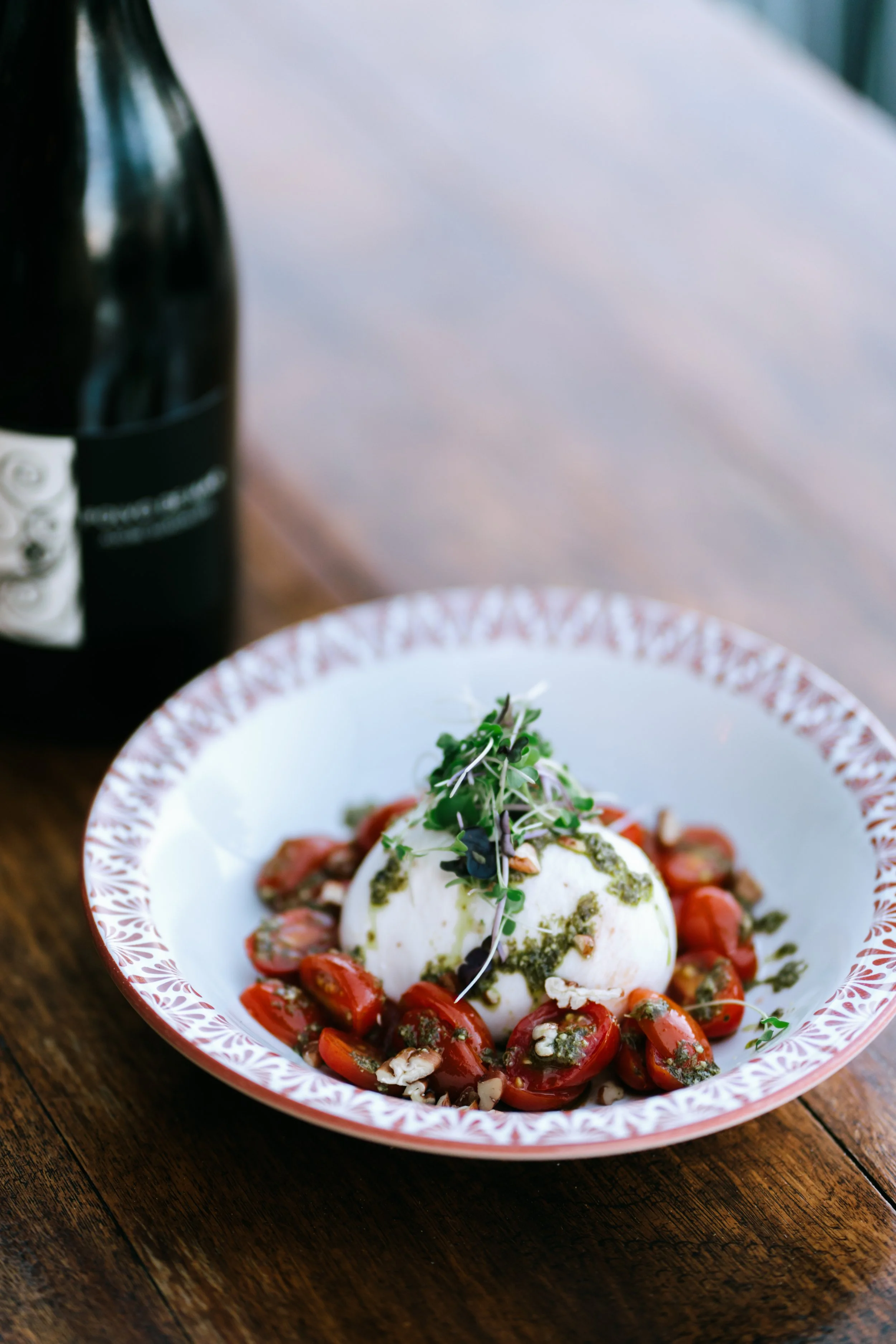 A plate of burrata cheese garnished with cherry tomatoes, herbs, and pesto, with a bottle of wine in the background.