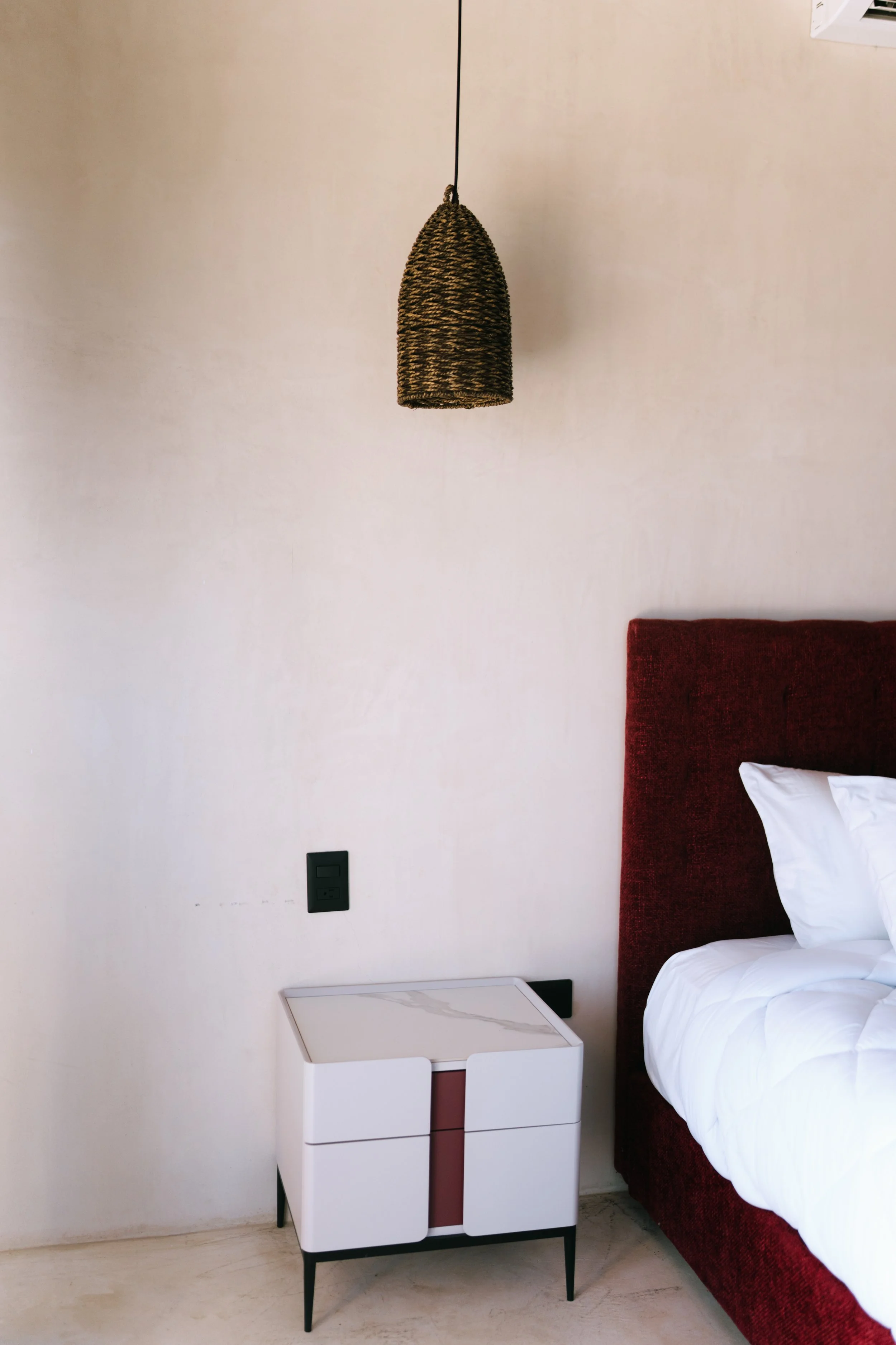 A modern bedroom corner featuring a white nightstand with a marble top, a red upholstered bed with white bedding, a woven pendant light hanging from the ceiling, and a wall air vent.