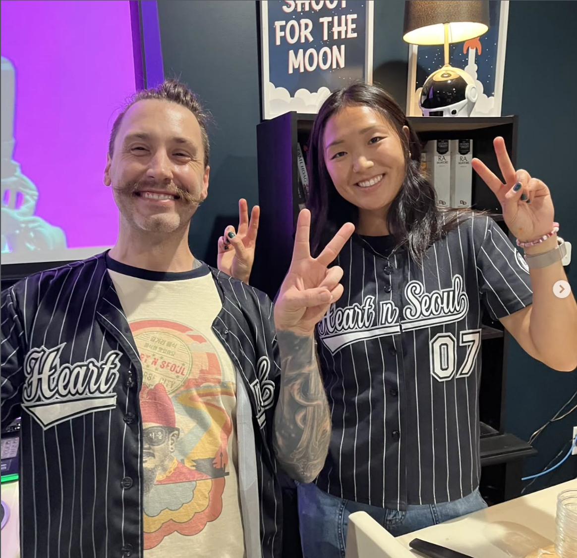 A couple in baseball jerseys emblazoned with Heart N Seoul flash the peace sign