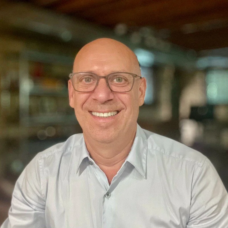 A smiling bald man wearing glasses and a white button-up shirt, indoors with shelves and a window in the background.