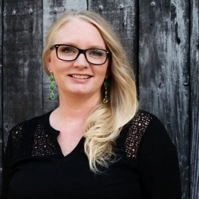 A woman with blonde hair, wearing glasses, green earrings, and a black top, standing in front of a weathered wooden fence.