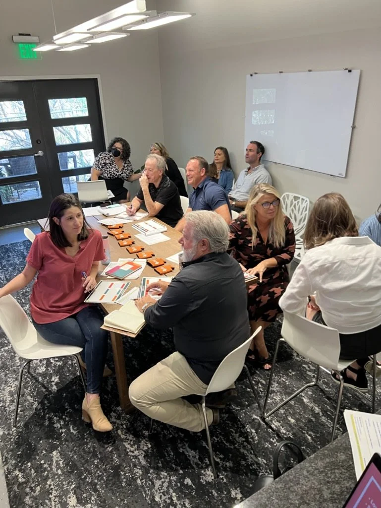 People in a meeting room seated around a table with documents and laptops, engaged in discussion.