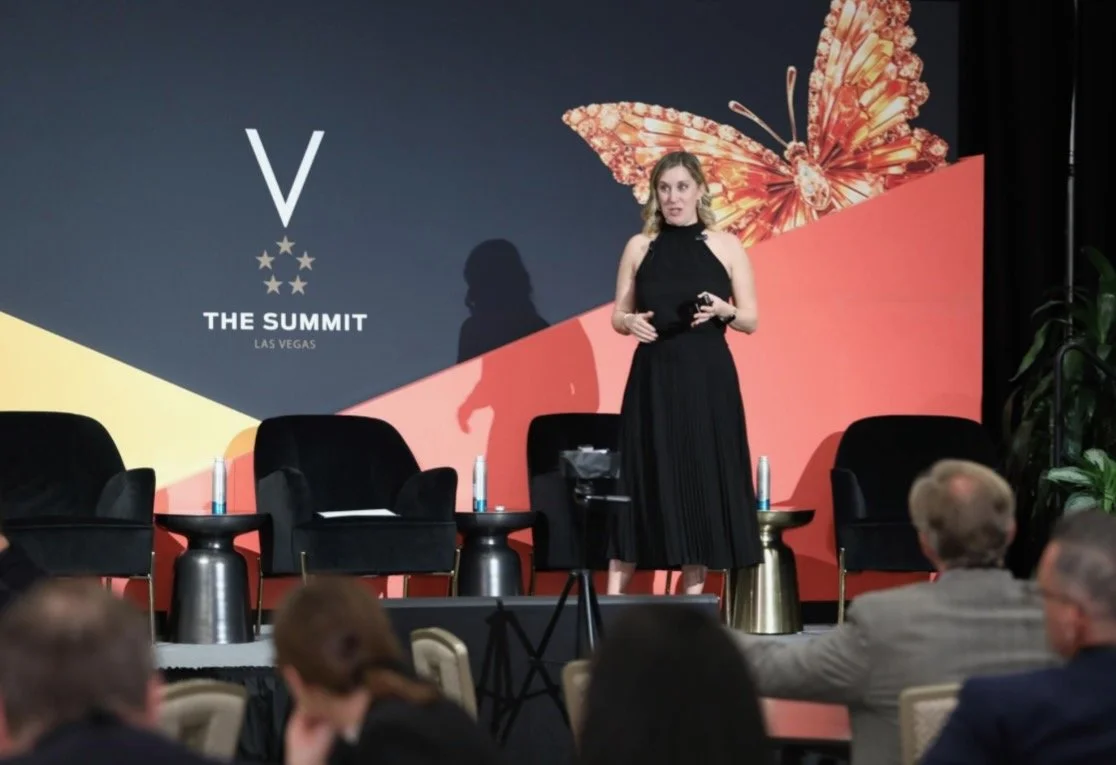 A woman in a black dress on stage speaking at a conference with a large butterfly graphic and the text 'The Summit Las Vegas' on a backdrop behind her, while audience members listen.
