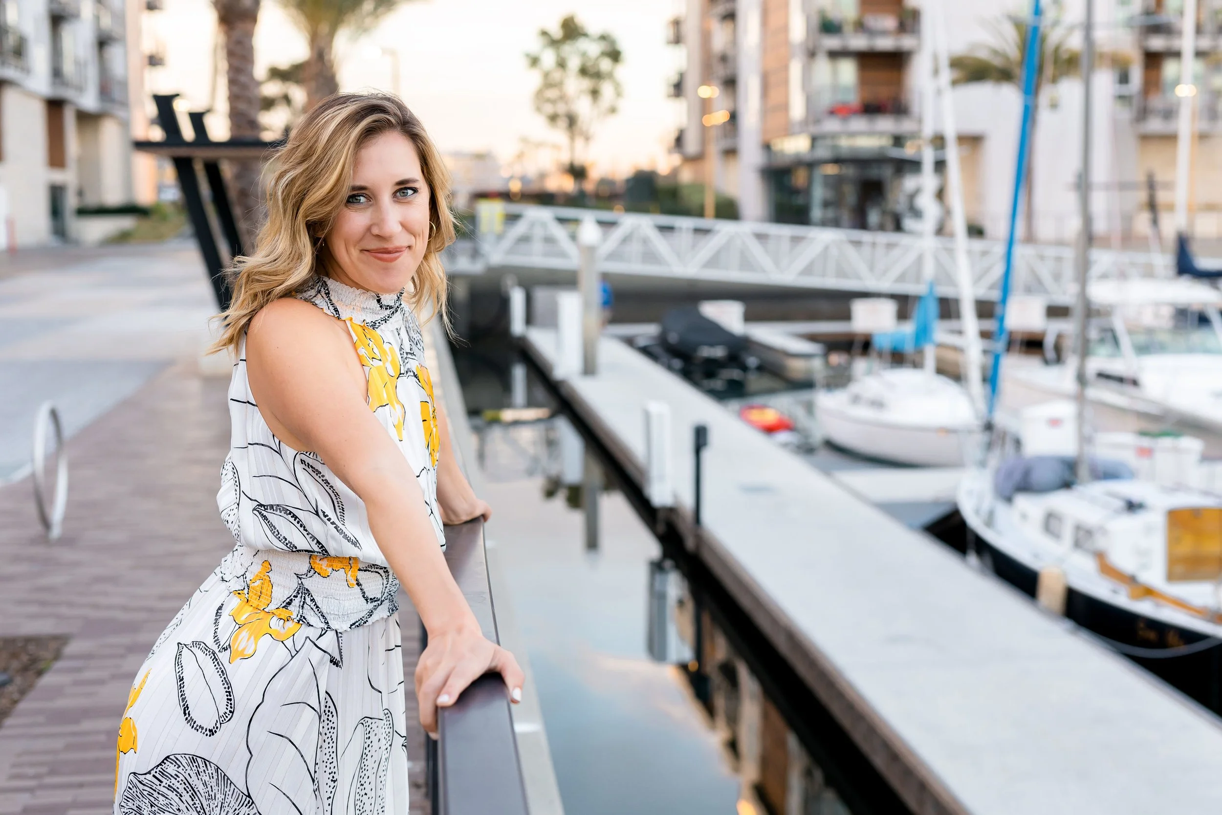 A woman with blonde hair smiling, standing on a walkway by a marina with boats docked, in the evening.