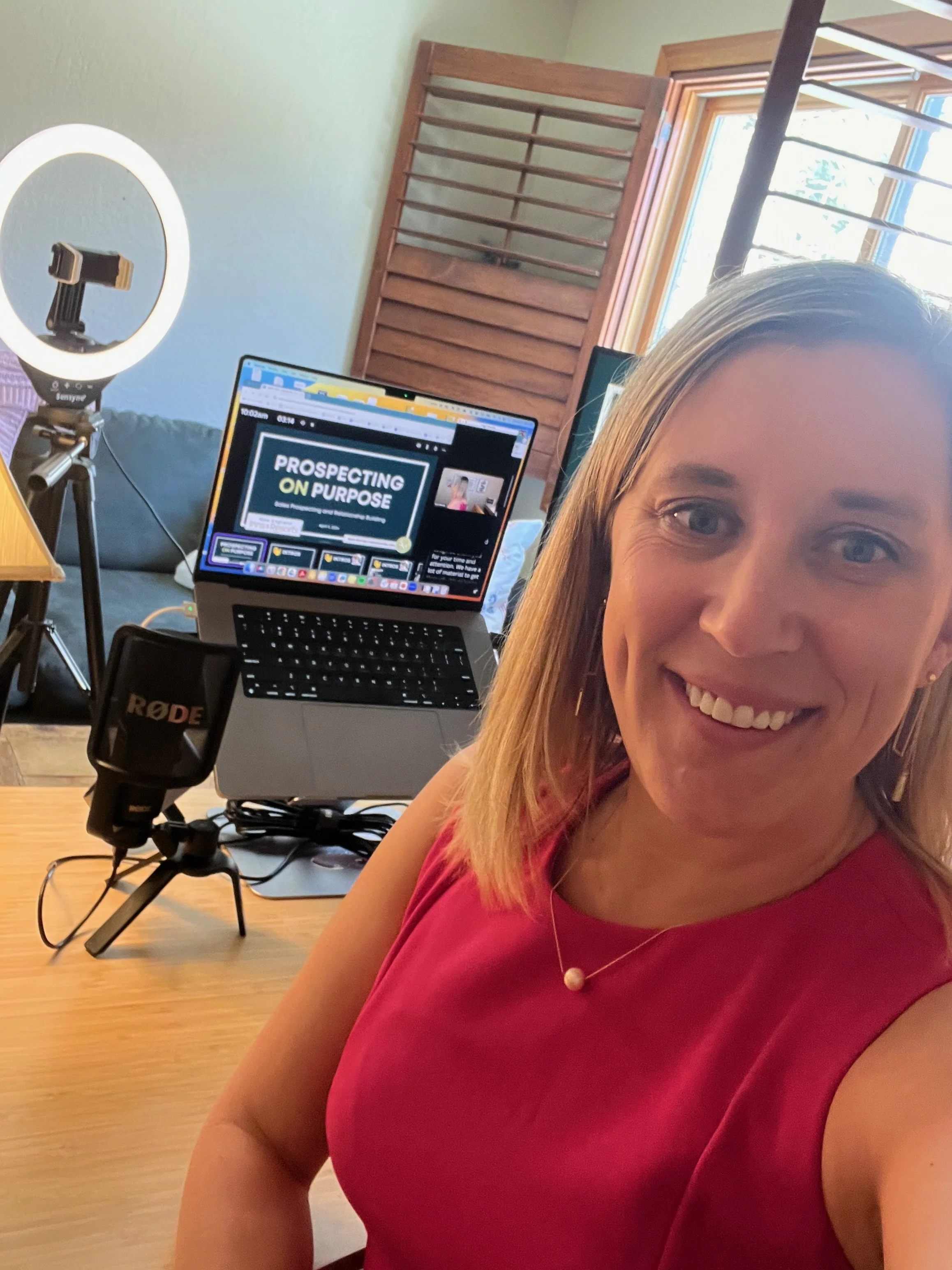 A woman taking a selfie in a home office setup with a ring light, a microphone, a laptop displaying a presentation titled 'Prospecting on Purpose,' and a window with sunlight streaming in.