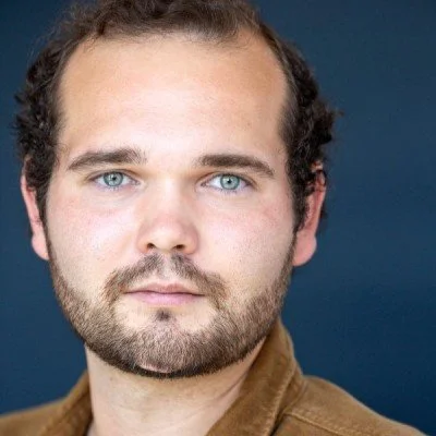 Close-up of a man with blue eyes, dark curly hair, and a beard, wearing a brown jacket against a dark background.