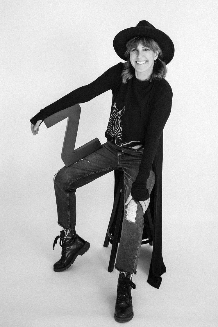 A smiling woman wearing a black wide-brim hat, black sweater with a zebra print, ripped jeans, and black boots, sitting on a stool and holding a large letter Z, against a plain background.