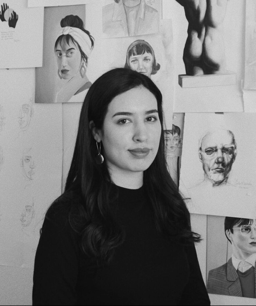 Black and white photo of a woman with long dark hair wearing a black top and earrings, standing in front of a wall covered with various portrait sketches and drawings.