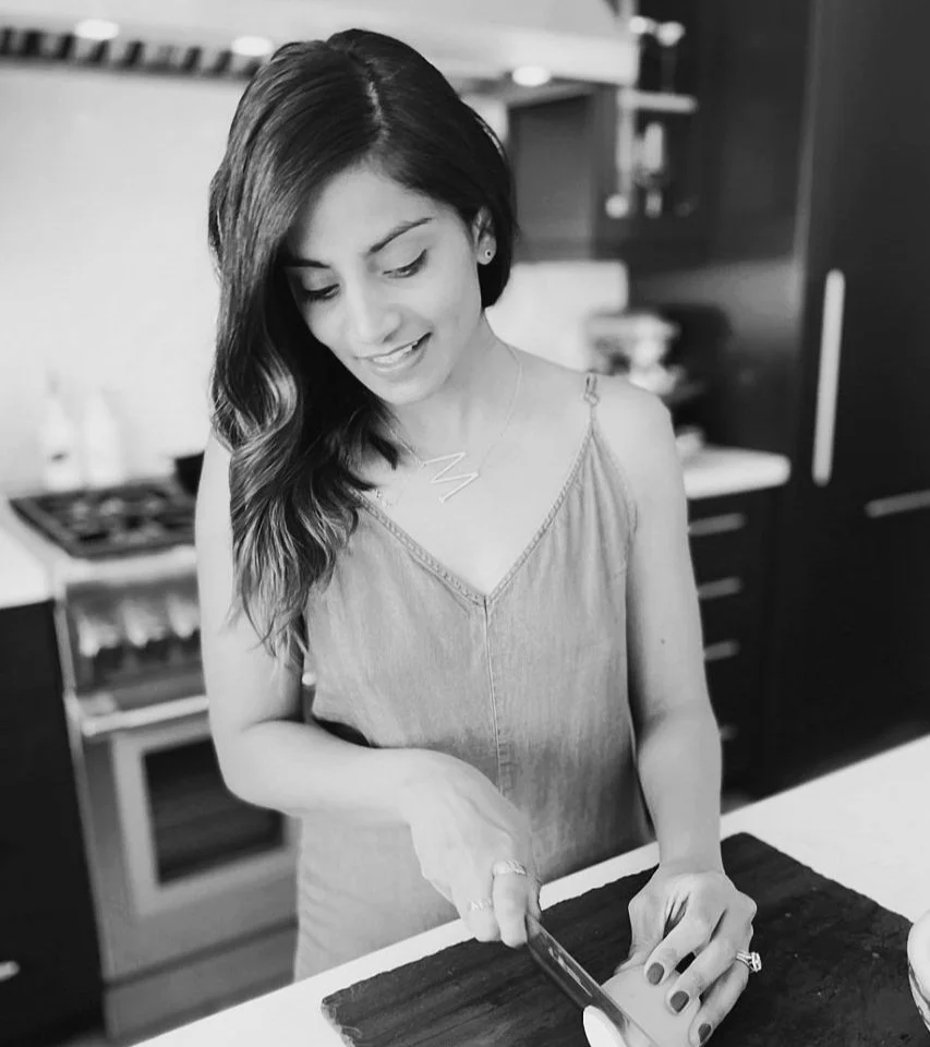 A woman with dark, wavy hair smiles while cutting something on a countertop in a kitchen.