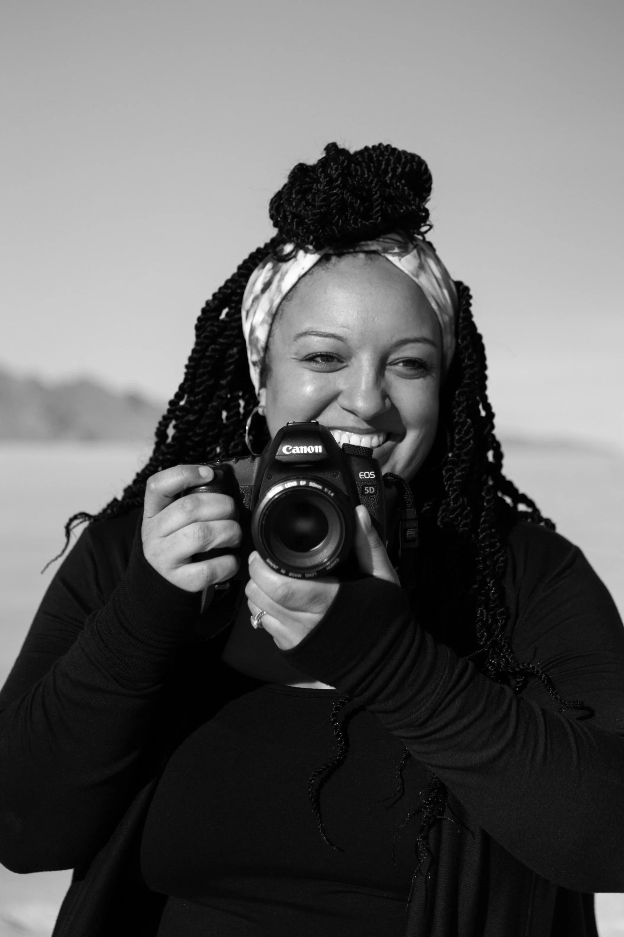 A smiling woman with twisted hair wearing a headscarf takes a photo with a Canon EOS camera in a bright outdoor setting.