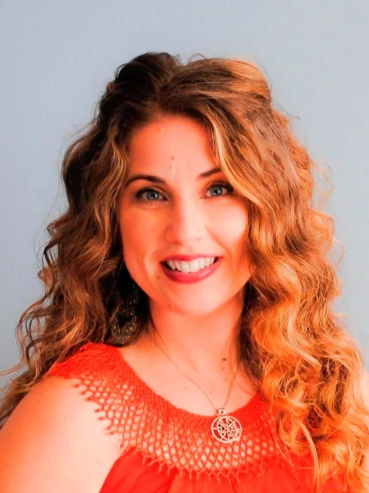 A woman with curly red hair wearing an orange top and a silver necklace with a pentacle pendant.