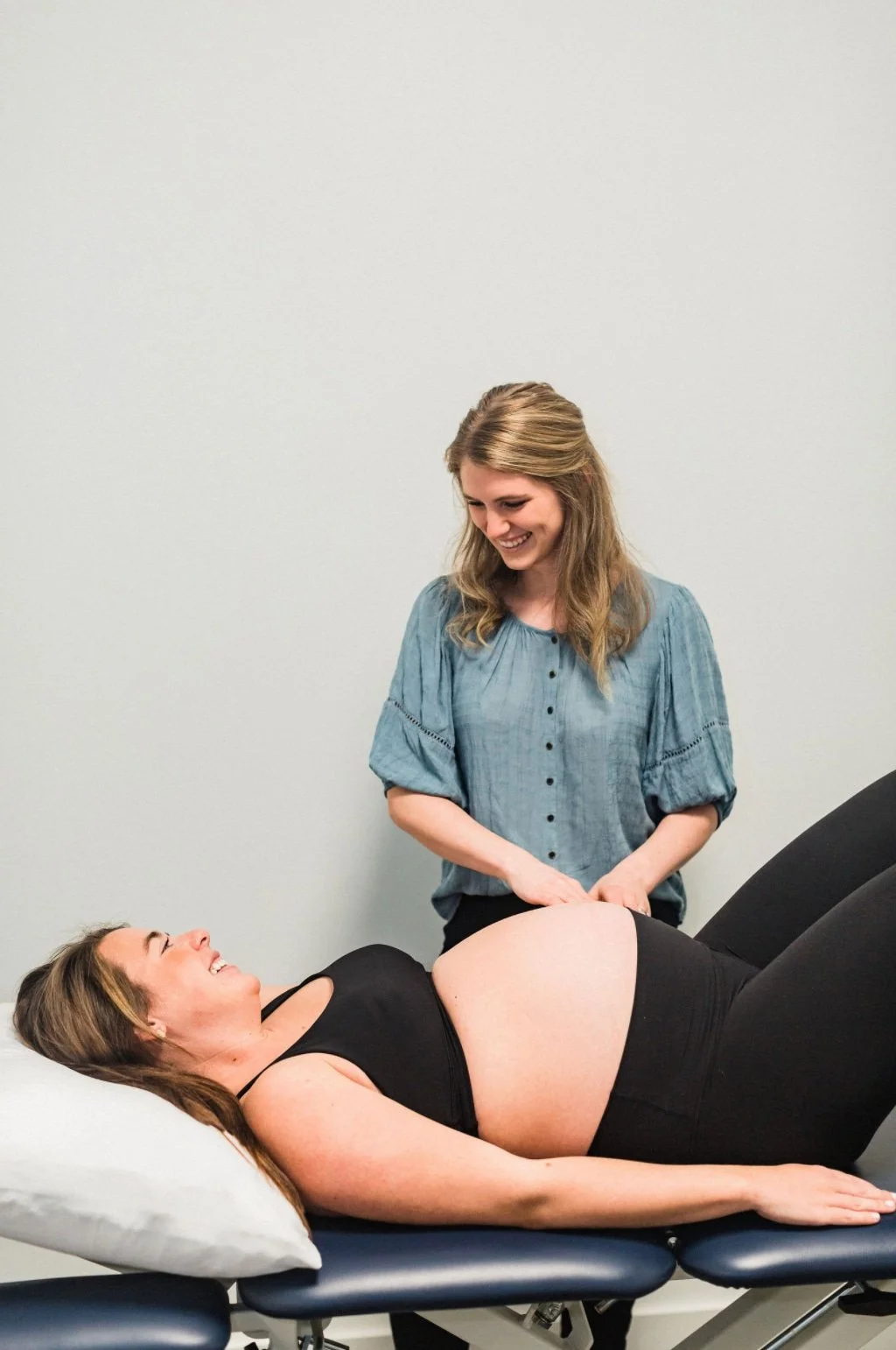 Pregnant Woman in black top and black bottoms laying on back. Women in Motion Physical therapist standing to patients left wearing green shirt, blonde hair and smiling