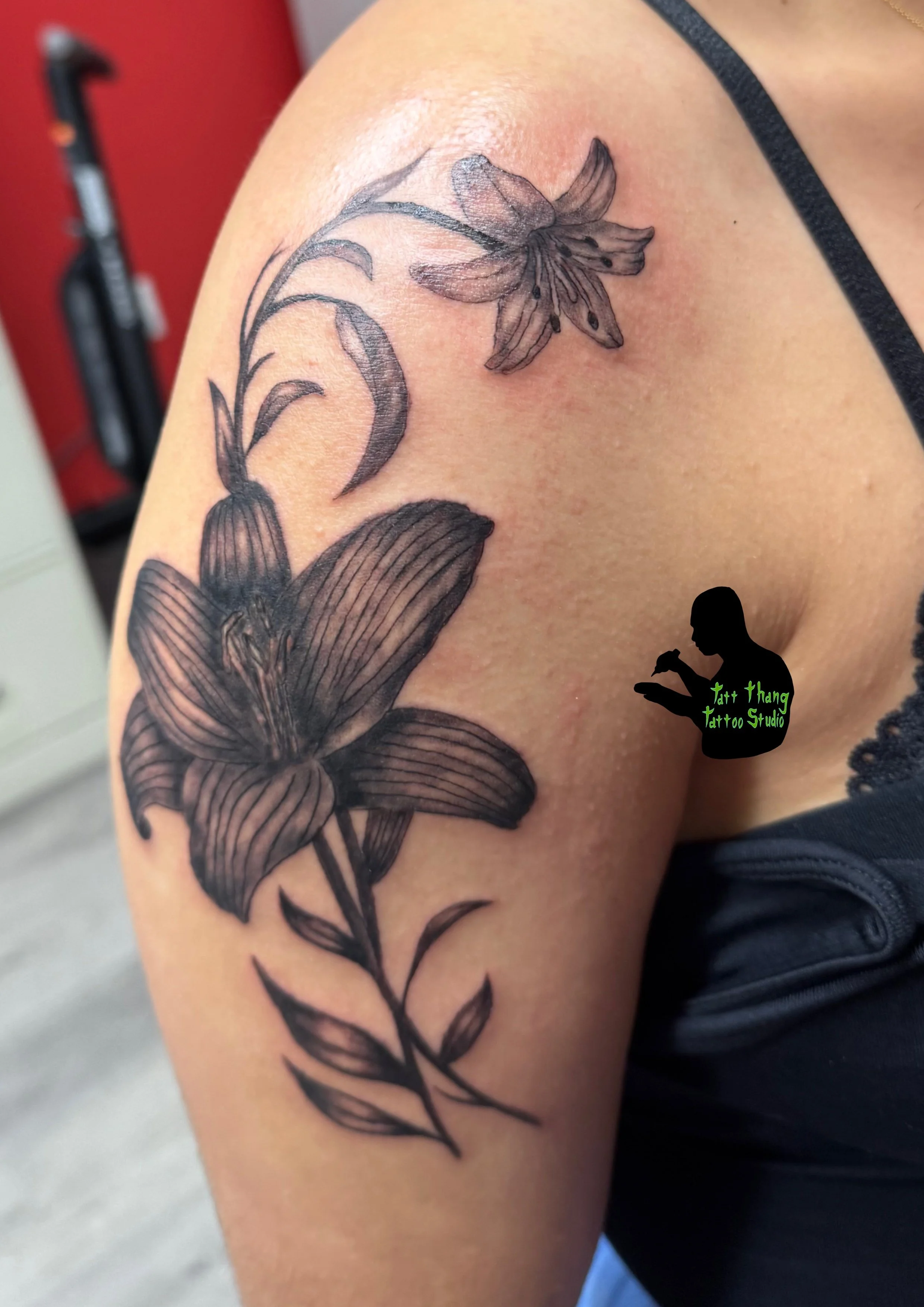 Black and gray Amaryllis tattoo on upper arm, with detailed petals, stem, and leaves.