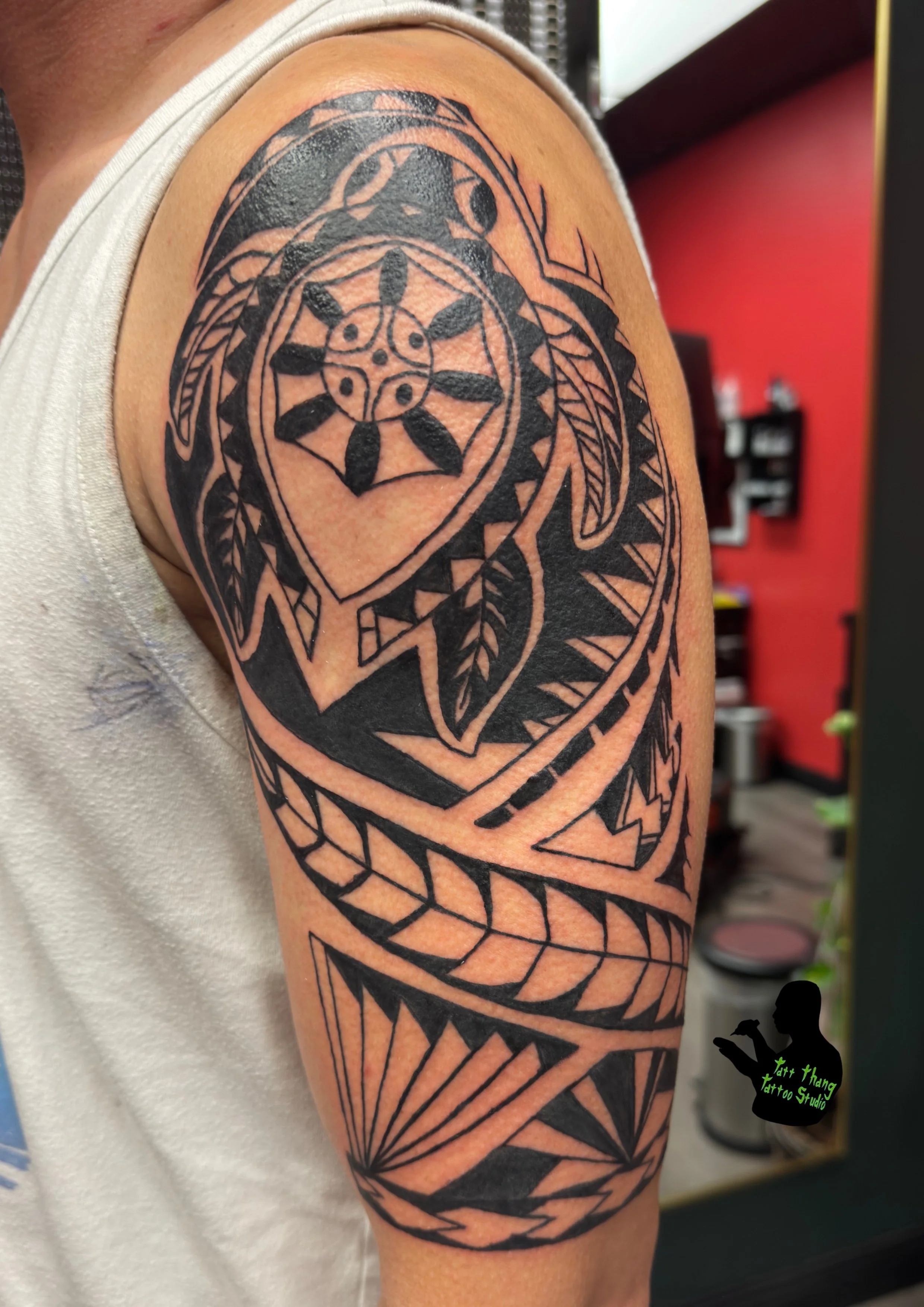 Black tribal-style tattoo on a person's upper arm featuring geometric shapes, a central circle with dots, leaves, and various patterns.
