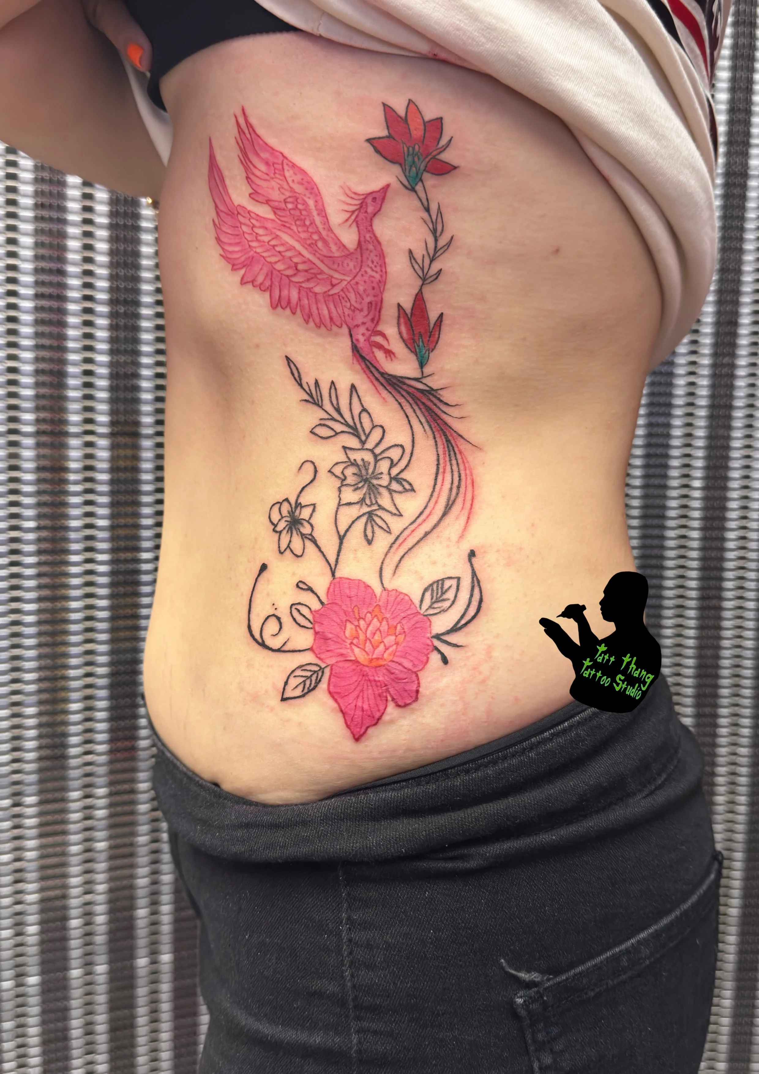 Colorful tattoo of a pink phoenix flying above a flower with detailed leaves and stems on a person's side.