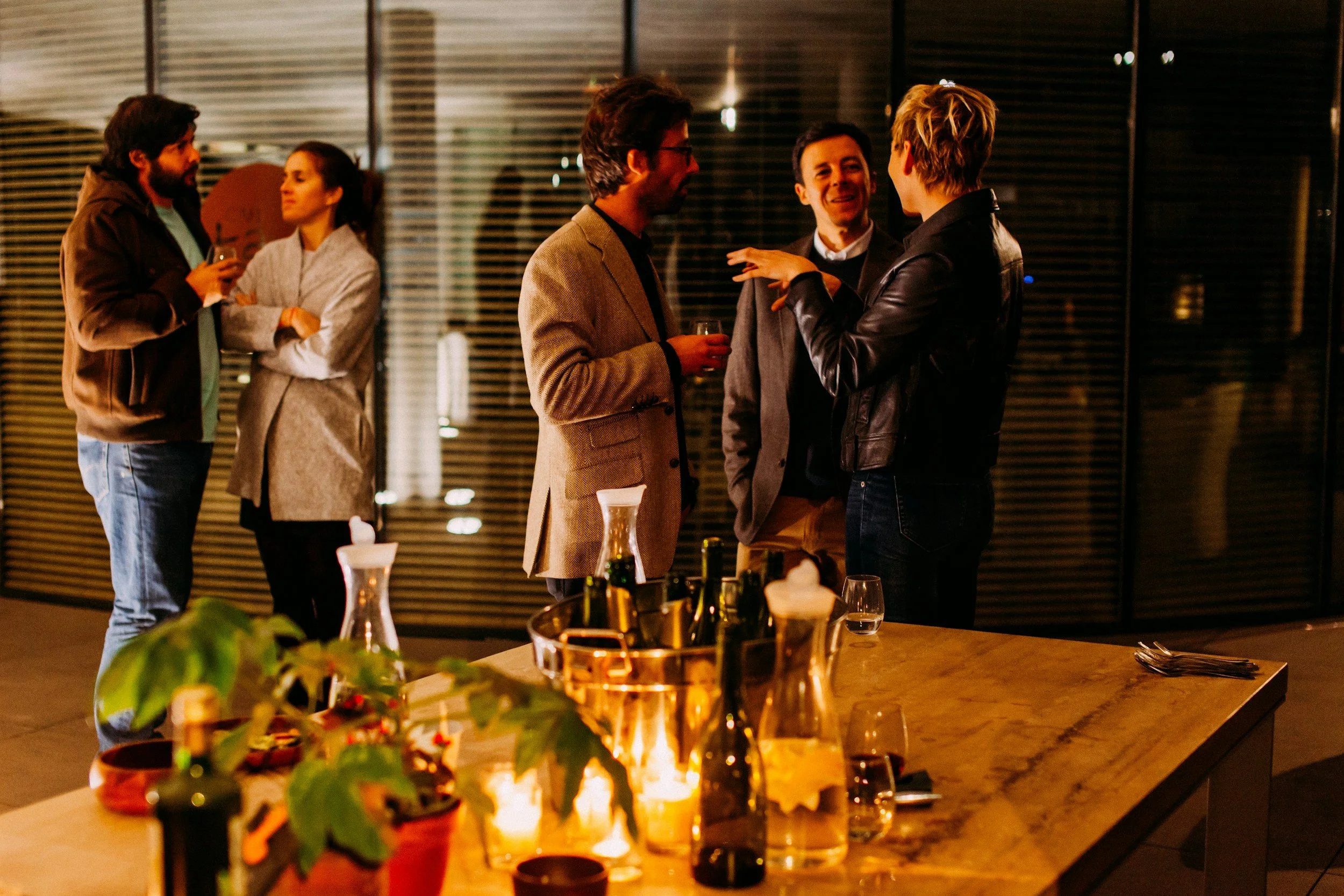 A group of six people socializing at a gathering, standing and talking in a warmly lit room with large windows or glass doors. A table with bottles, glasses, and a potted plant is in the foreground.