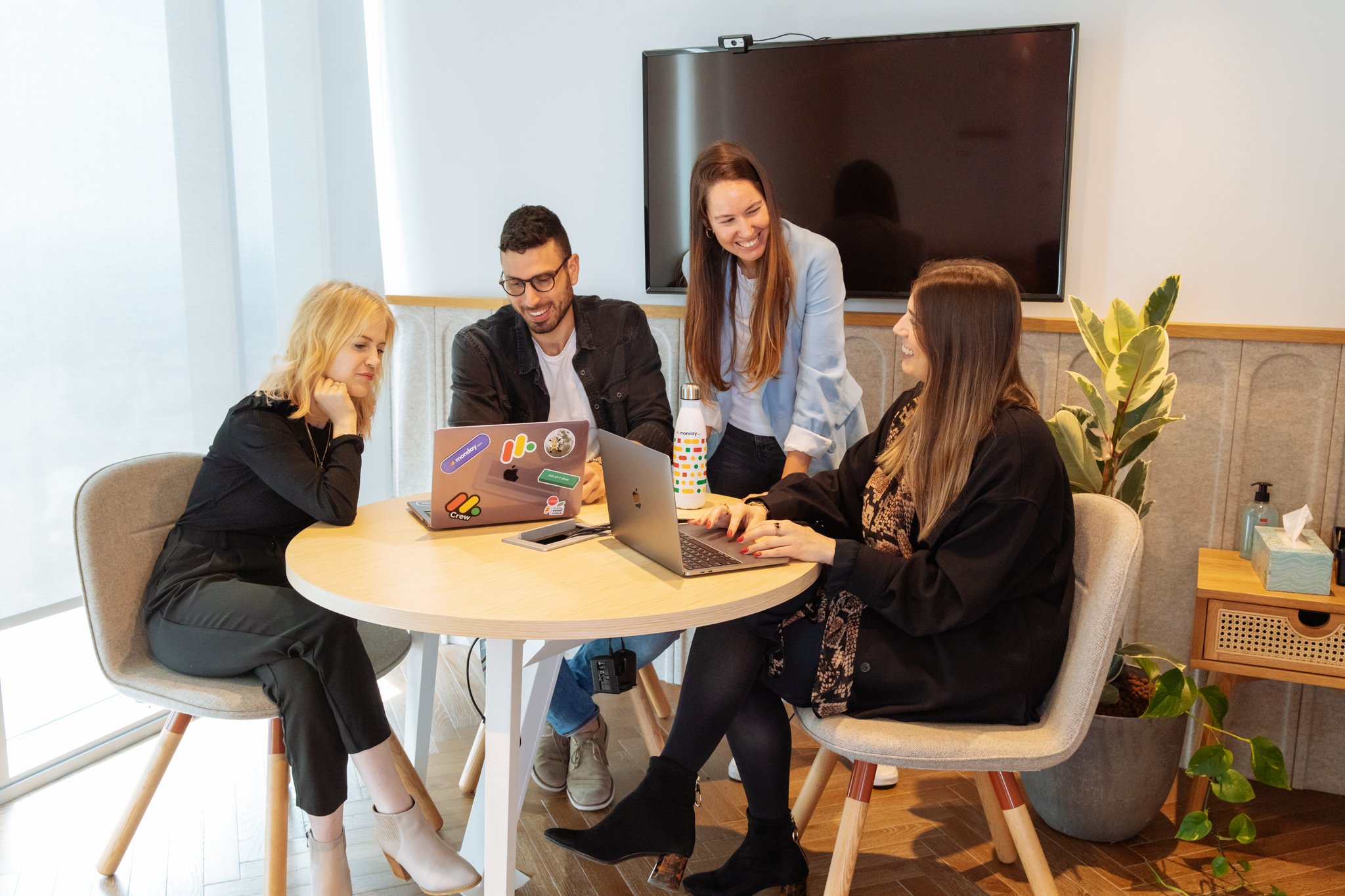 Five young professionals smiling and working together at a round table in a modern office, with laptops, a water bottle, and a TV screen on the wall.