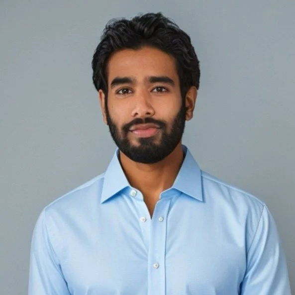 Portrait of a man with dark hair and beard, wearing a light blue collared shirt, against a gray background.