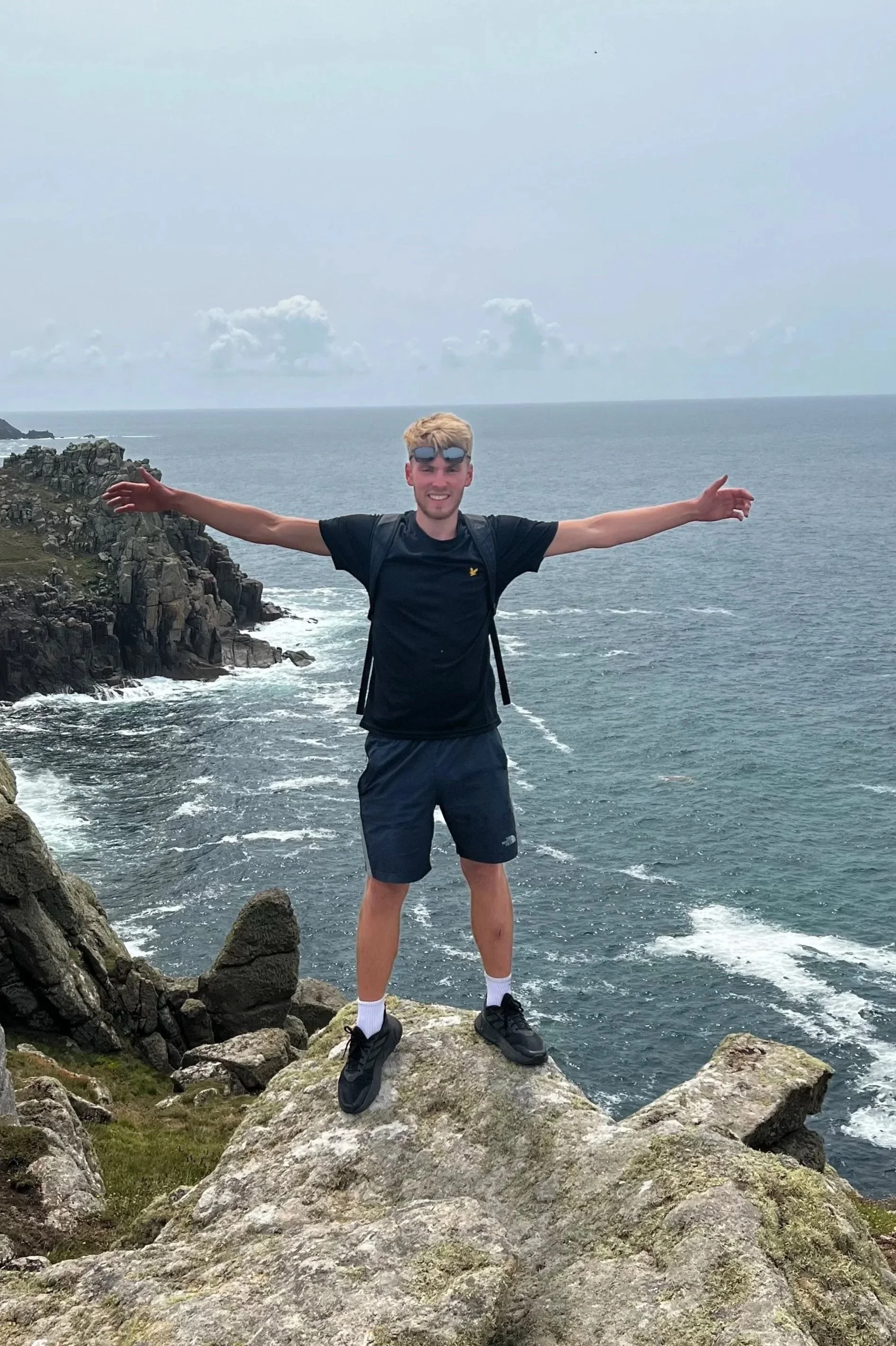 A young man standing on a large rock with arms outstretched in front of a rocky coastline and ocean under a cloudy sky.