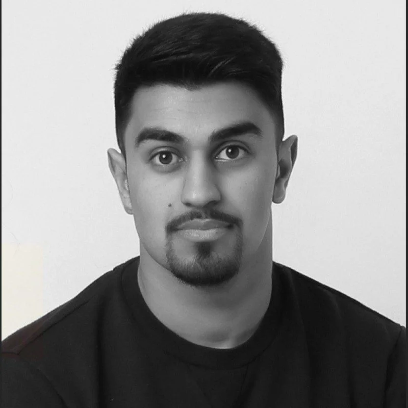 A black-and-white portrait of a young man with short black hair, a goatee, and wearing a black shirt, looking directly at the camera against a plain white background.