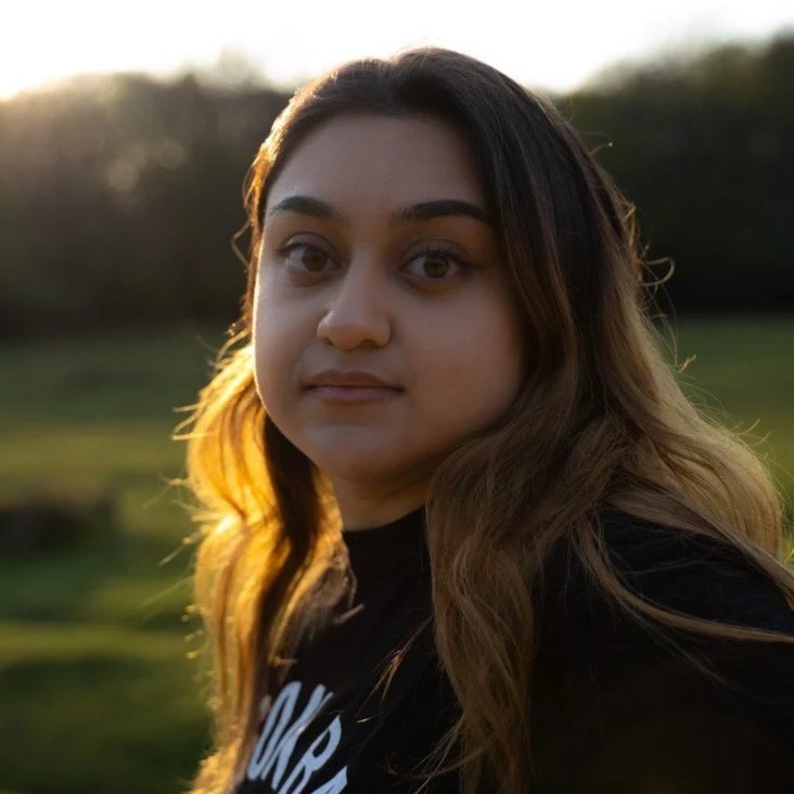 A young woman with long, wavy hair and natural makeup outdoors during sunset, with a blurred green background.