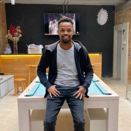 A man sitting on a pool table in an indoor room, smiling at the camera with a TV mounted on the wall behind him and a floral decoration on a table to the left.