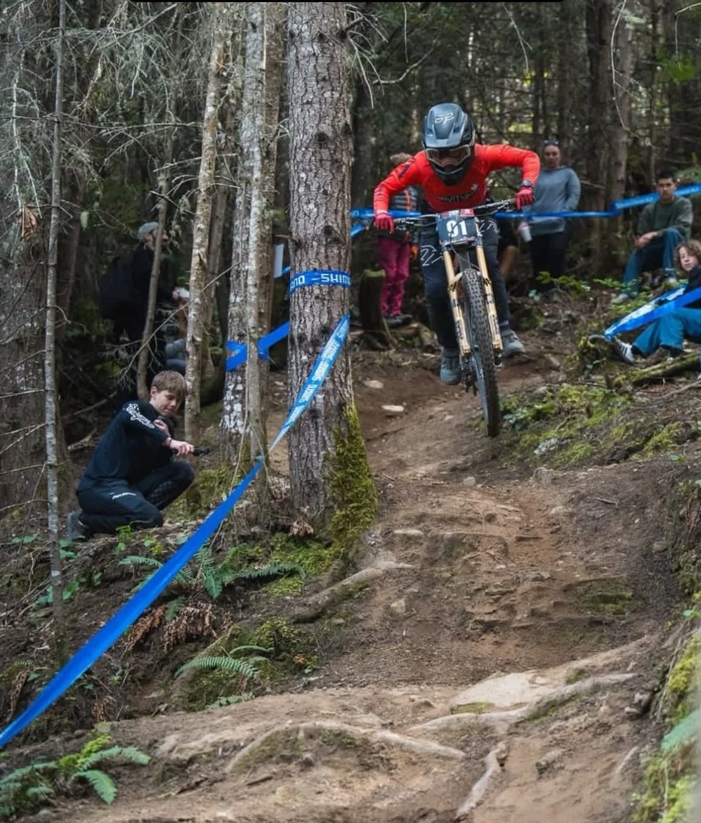 A mountain biker wearing a red jersey, helmet, and goggles jumping over a dirt trail in a forest during a race, with spectators watching from the sidelines.