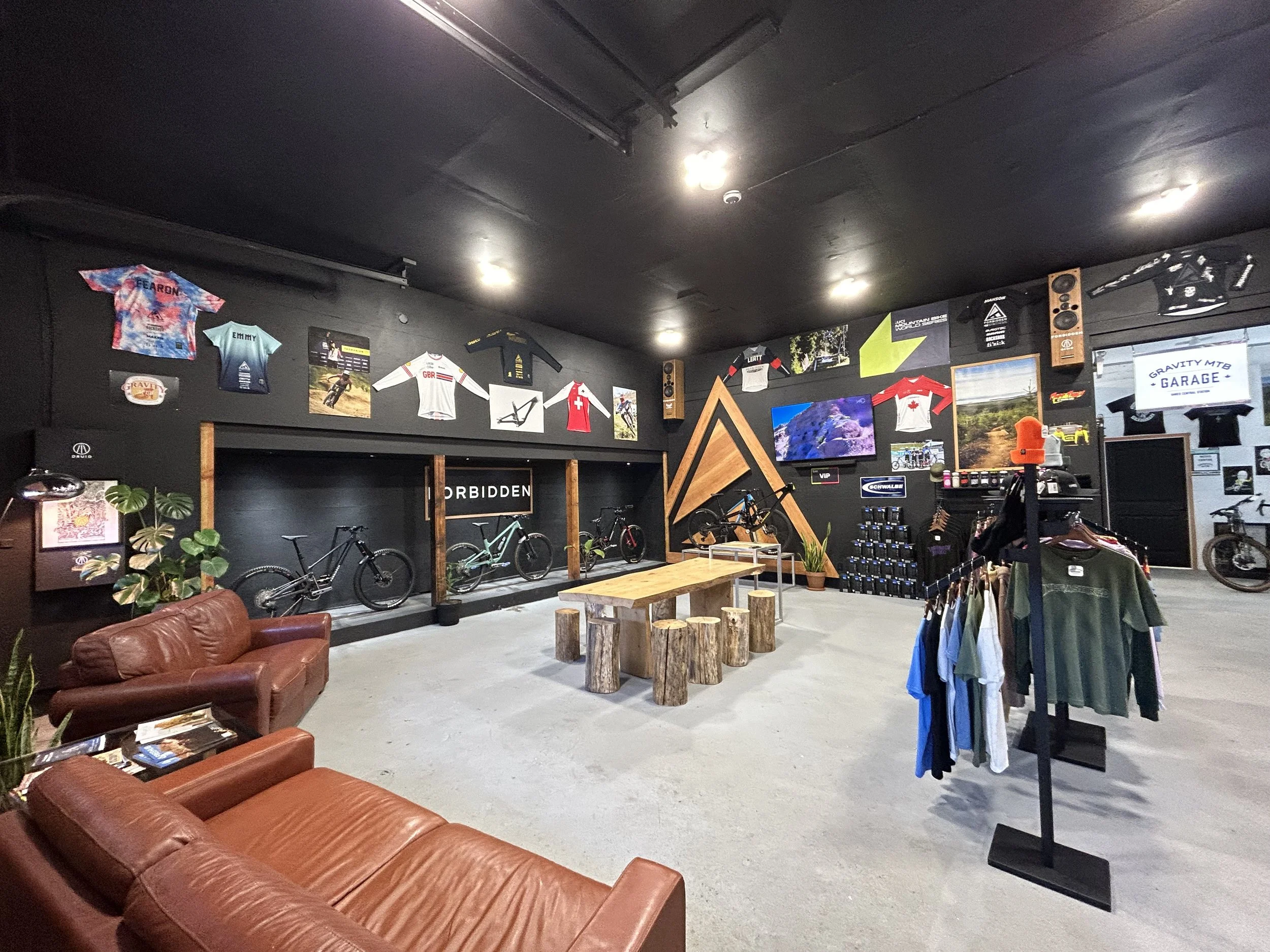 Interior of a bike shop with bikes, apparel, and memorabilia on the walls, leather couches, a wooden table with stool seats, and a flat-screen TV.