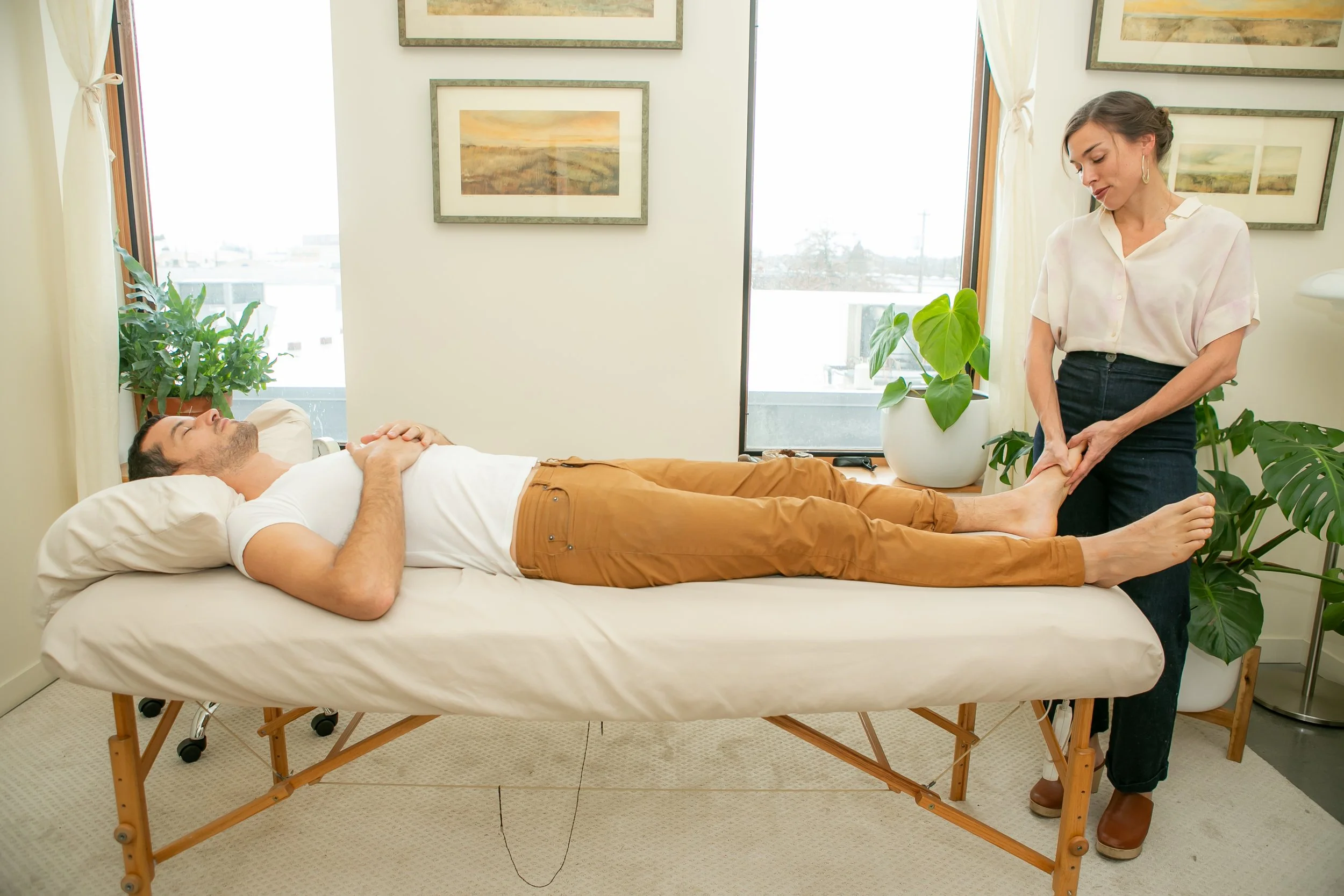 A woman giving a foot massage to a man lying on a massage table in a room with large windows and potted plants.
